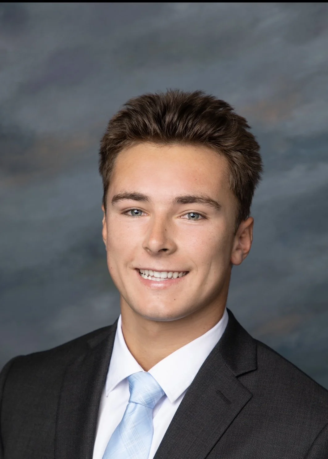 A young man smiling in a professional portrait, wearing a black suit, white shirt, and light blue tie, with a blurred cloudy sky background.