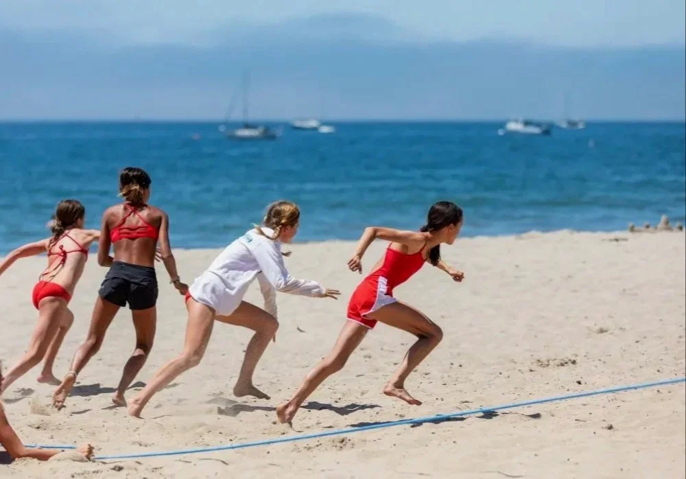 Four girls on a sandy beach in swimsuits and casual clothing, running towards the ocean with sailboats visible in the distance.