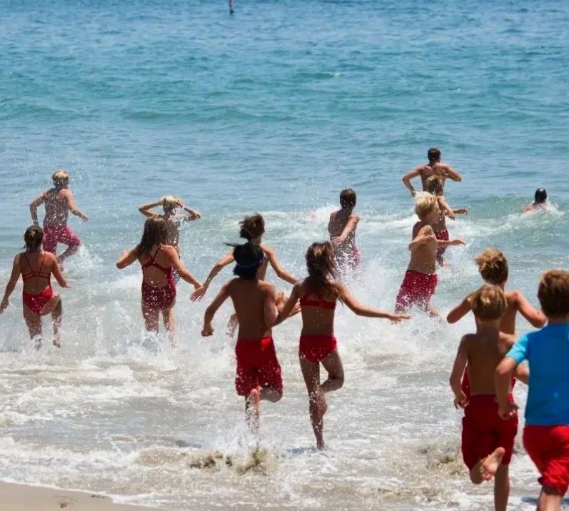 Children playing and running into the ocean at a beach, wearing swimsuits, with waves splashing around them.