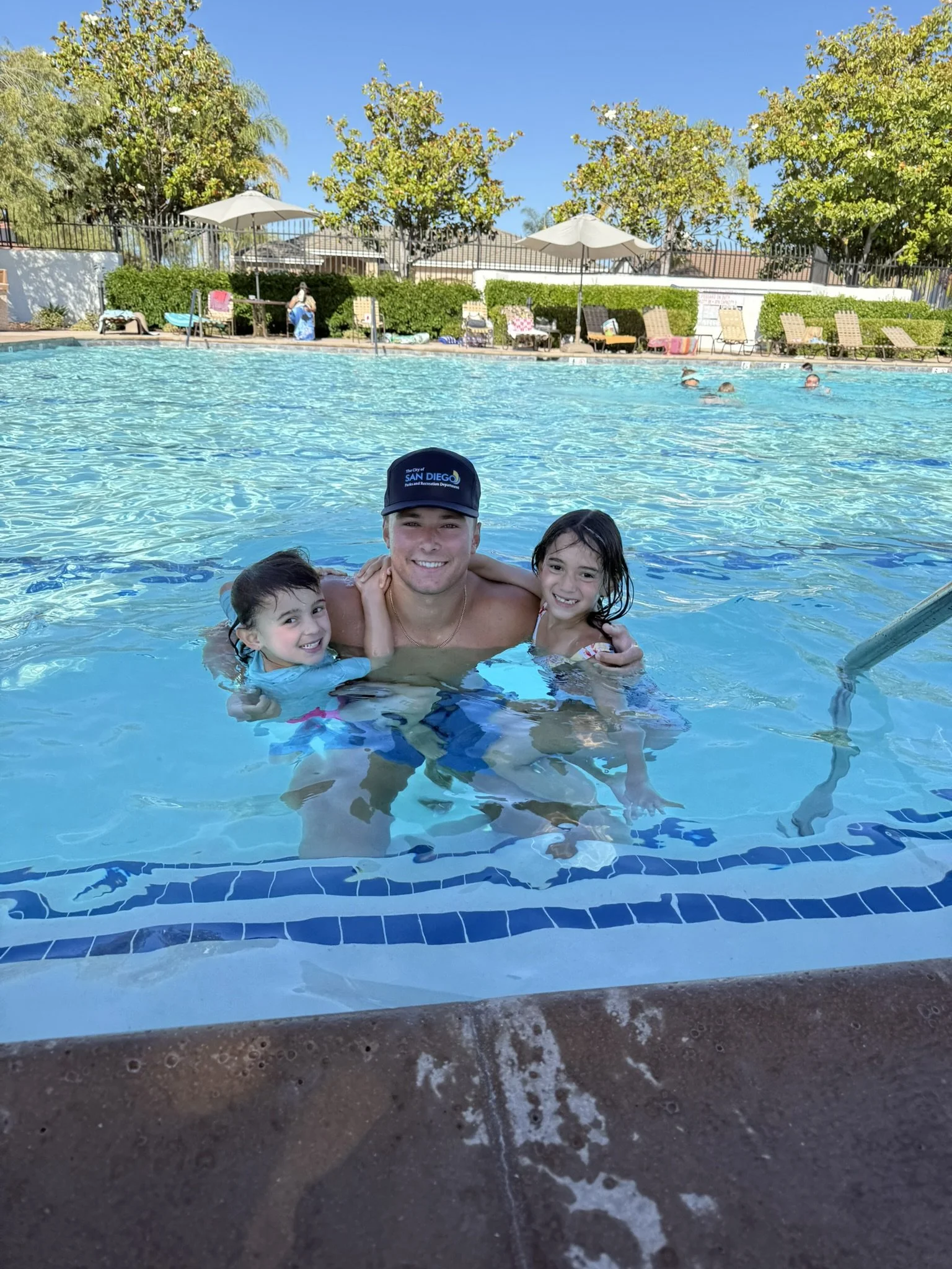 Three children and an adult man smiling and enjoying themselves in an outdoor swimming pool on a sunny day, with lounge chairs, umbrellas, and trees in the background.