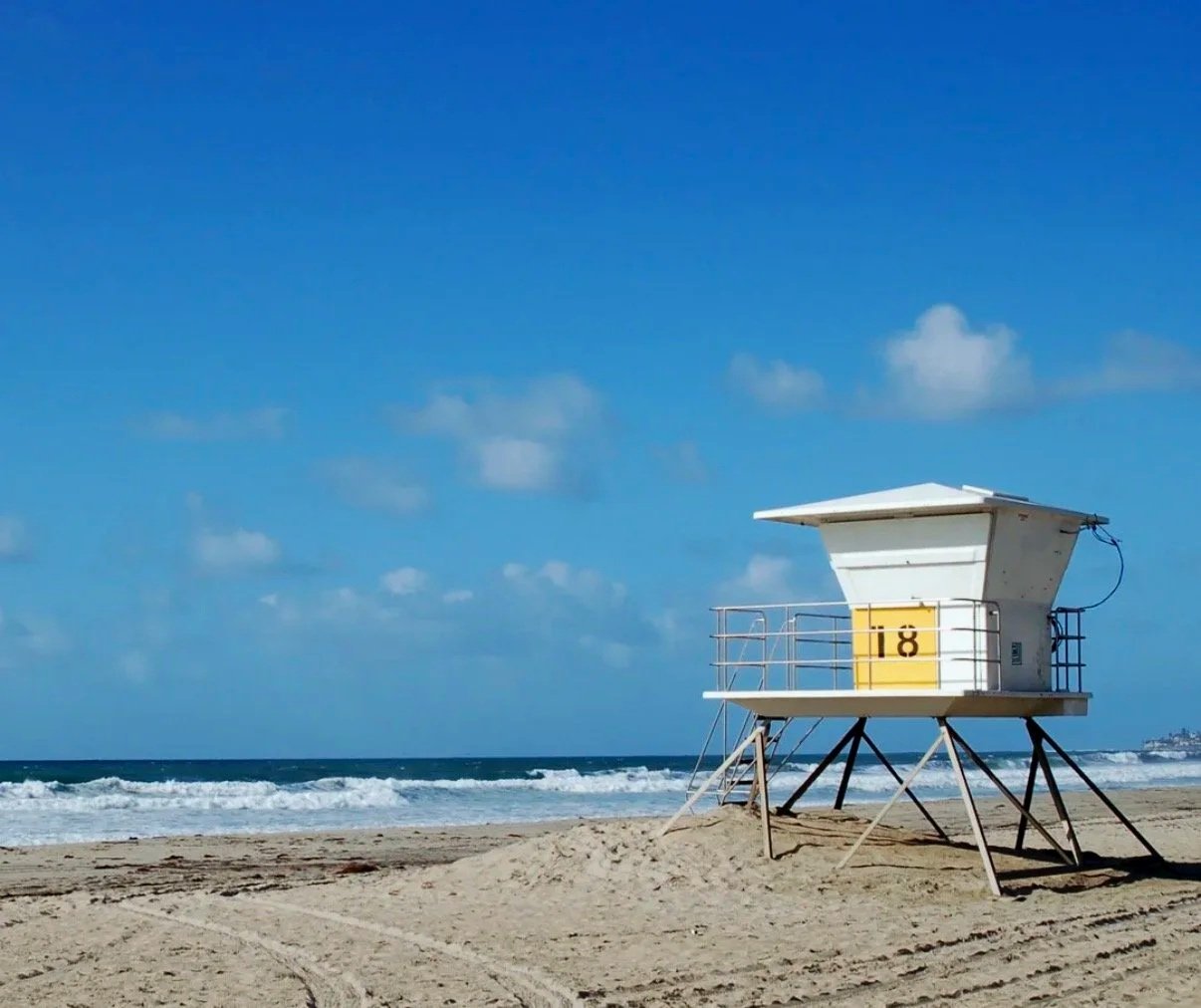 A lifeguard tower on a sandy beach with the ocean and blue sky with clouds in the background.