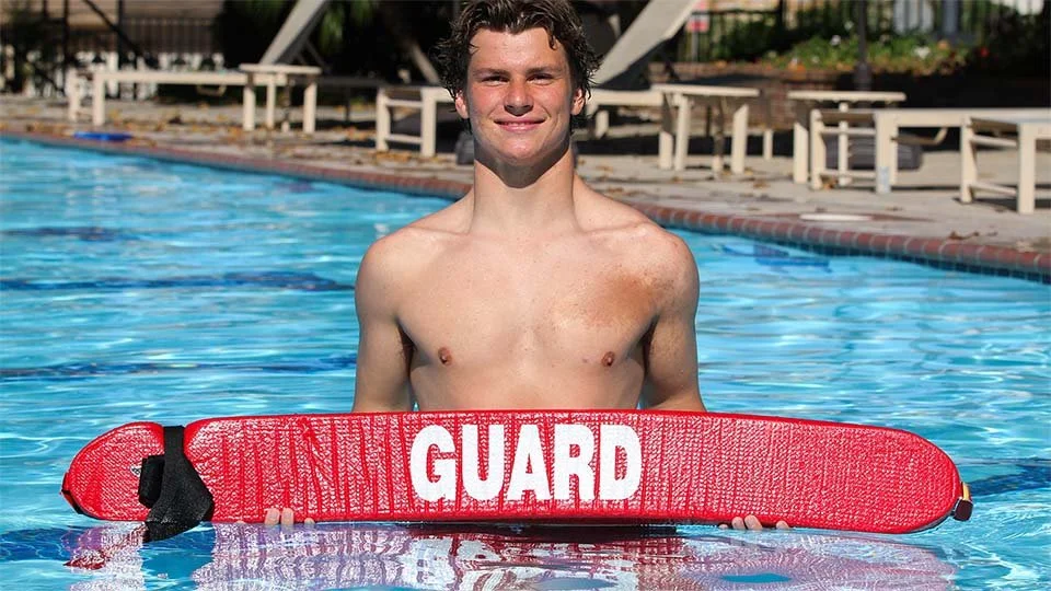 A young man standing in a swimming pool holding a red lifeguard board with the word 'GUARD' on it, smiling at the camera.