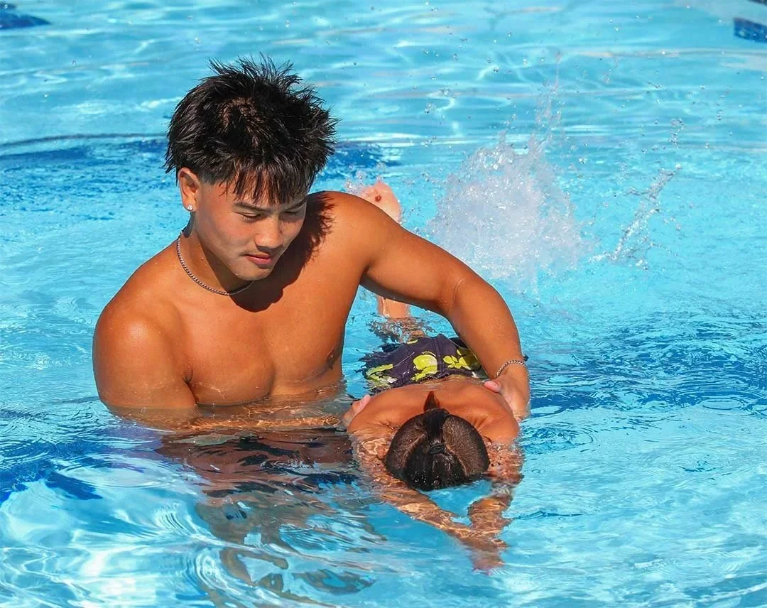 A young man is assisting a young girl with swimming in a bright blue pool. The man is shirtless, and the girl is wearing a swimsuit. The man is holding the girl as she floats face down in the water.