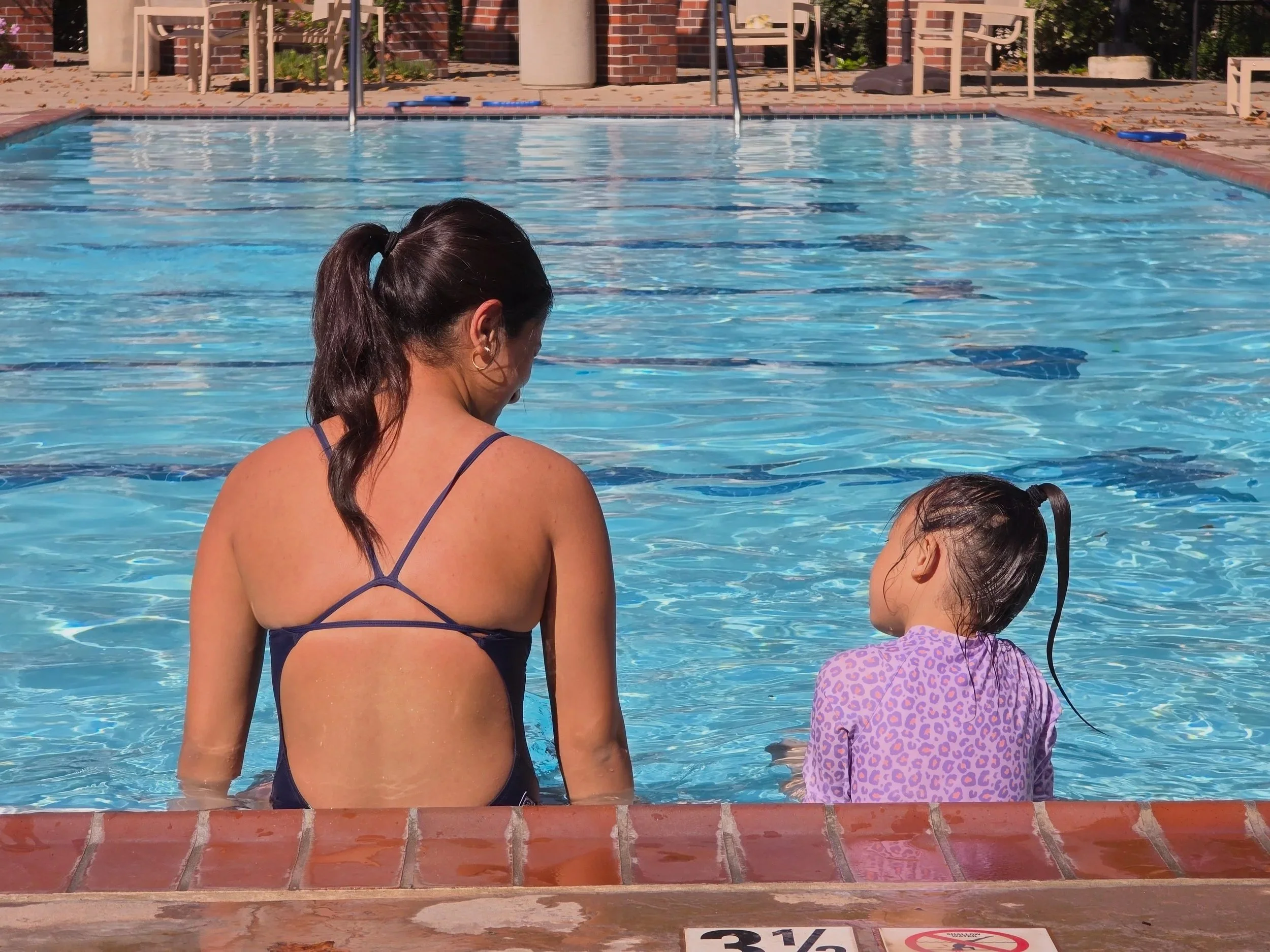 A woman and a girl standing at the edge of a swimming pool, with the woman observing the water and the girl close to the pool's edge. The pool has clear blue water with lane markings, and there are chairs and tables in the background.