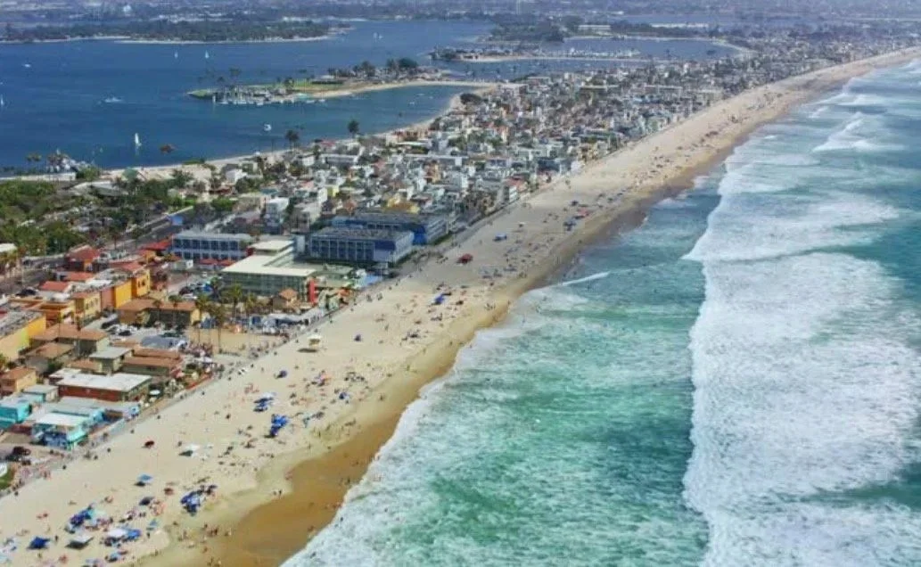 Aerial view of a beach with waves, sand, and beachgoers, alongside a town with buildings and streets, near a body of water with a marina or harbor in the background.
