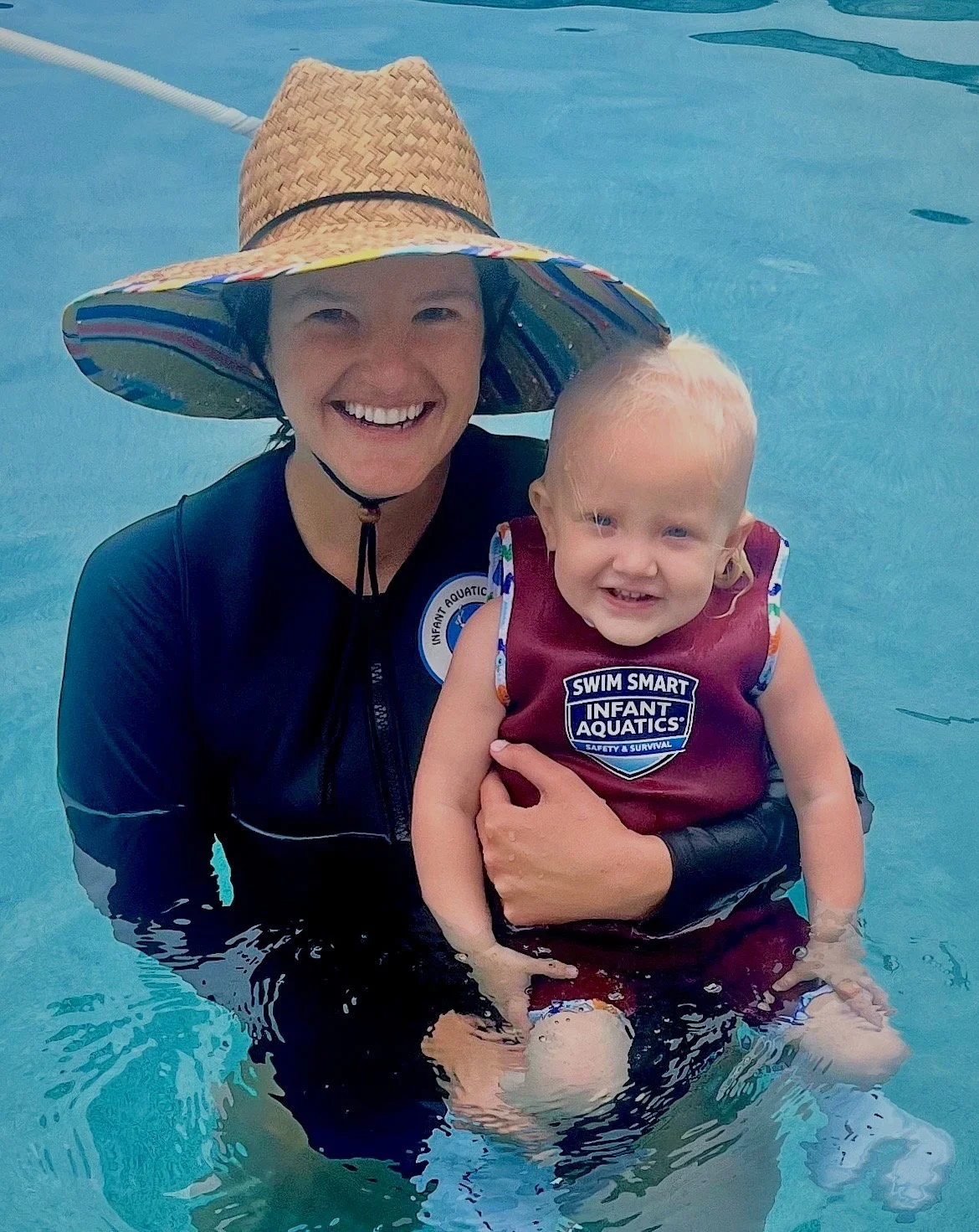 A woman and a young child smiling in a swimming pool. The woman is wearing a wide-brimmed straw hat and a dark long-sleeve swimsuit, holding the child. The child is wearing a red life jacket with glasses and has short blonde hair.
