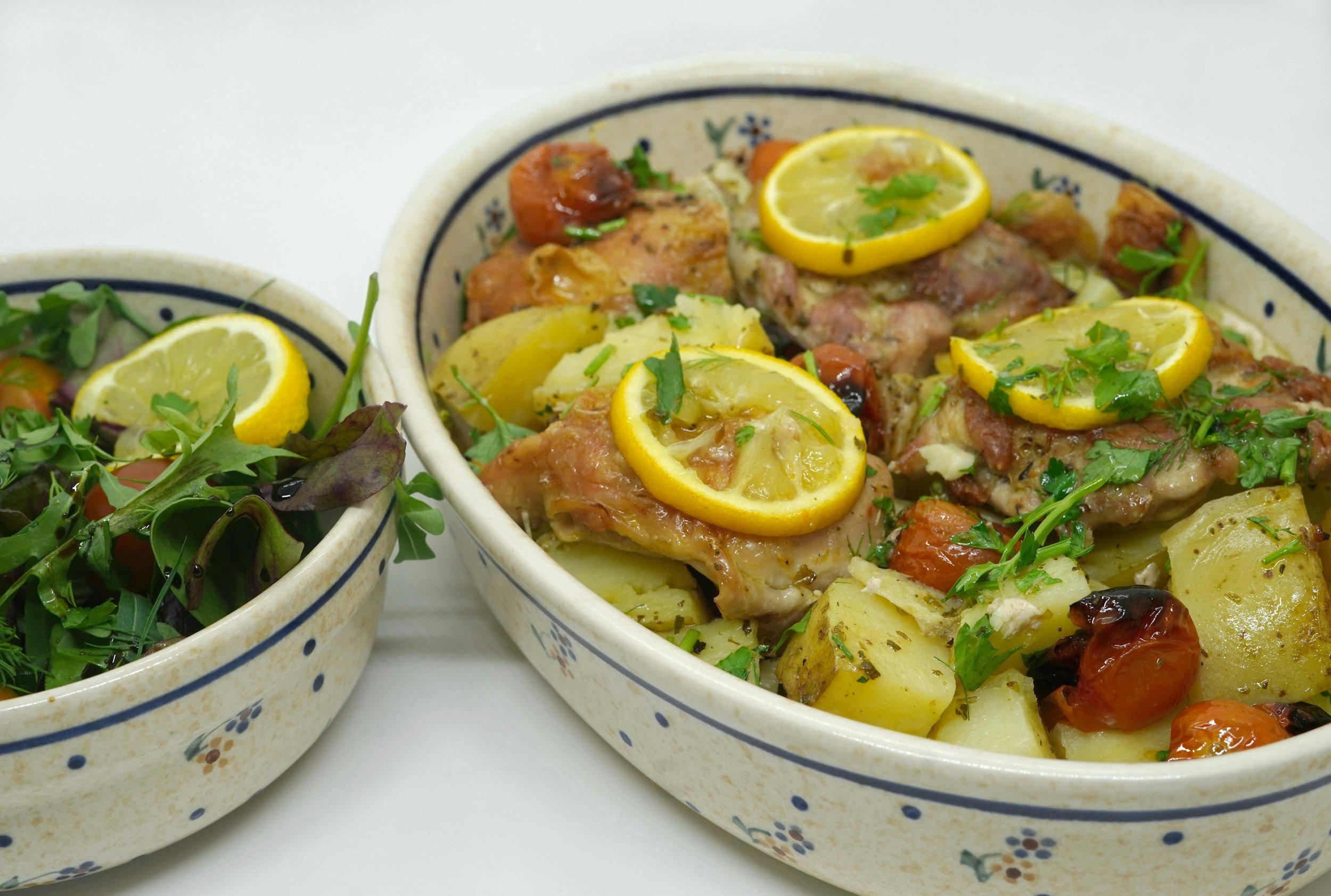 Two ceramic bowls with a blue and white floral design, filled with roasted chicken, potatoes, cherry tomatoes, and garnished with lemon slices and fresh herbs.