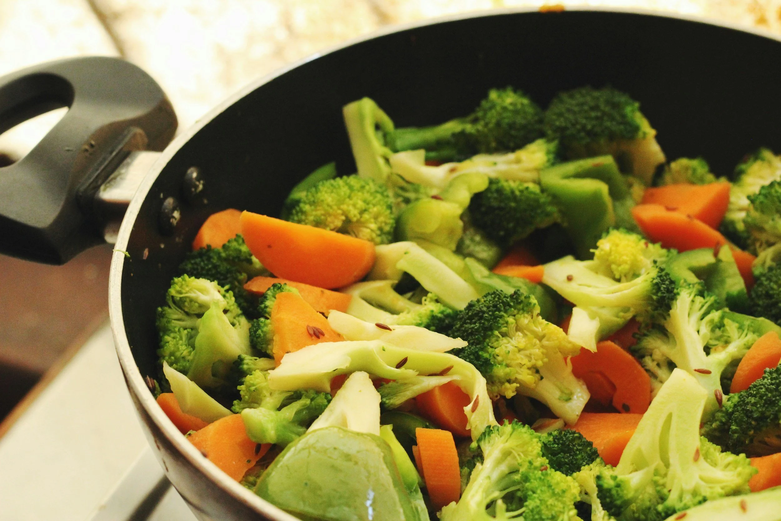 A black frying pan filled with steaming cooked broccoli, carrots, and cabbage on a stovetop.
