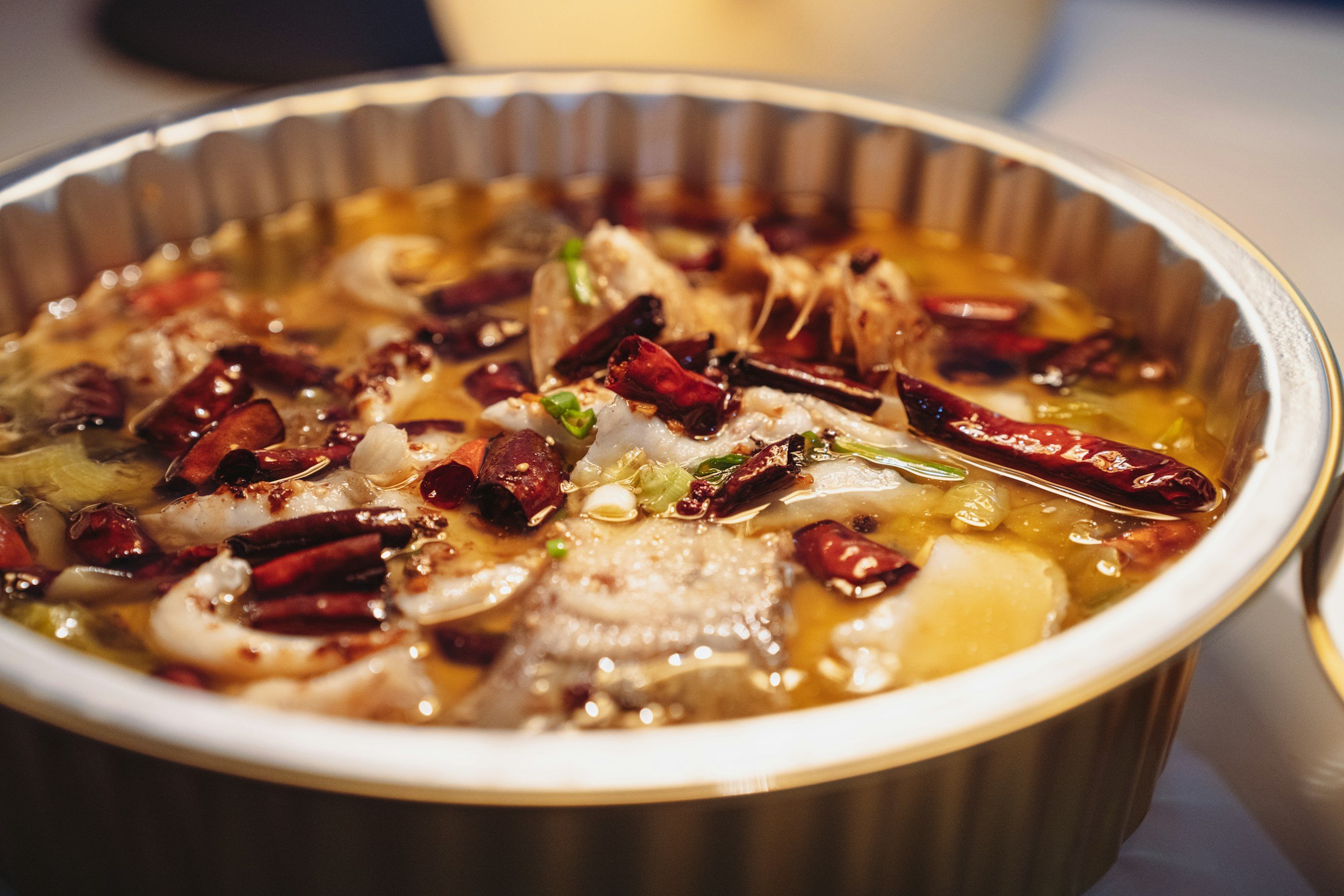 Close-up of a cooking pot filled with a spicy broth containing dried red chili peppers, chopped green onions, and pieces of meat.