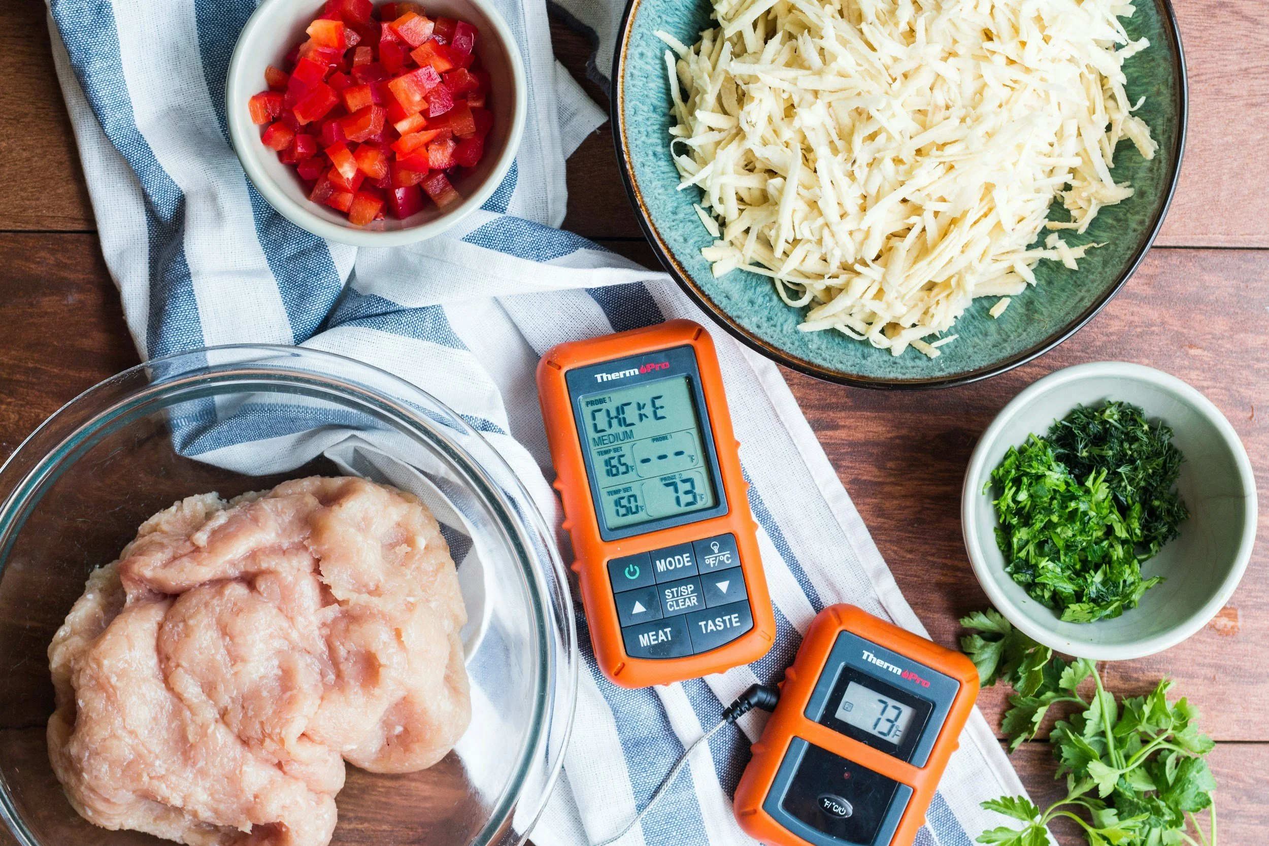 A collection of cooking ingredients including raw chicken in a glass bowl, shredded cheese in a green bowl, chopped tomatoes in a white bowl, chopped herbs in a small bowl, and digital thermometers measuring the temperature of the chicken at 165°F and 73°F, all arranged on a wooden surface with a blue and white striped cloth.
