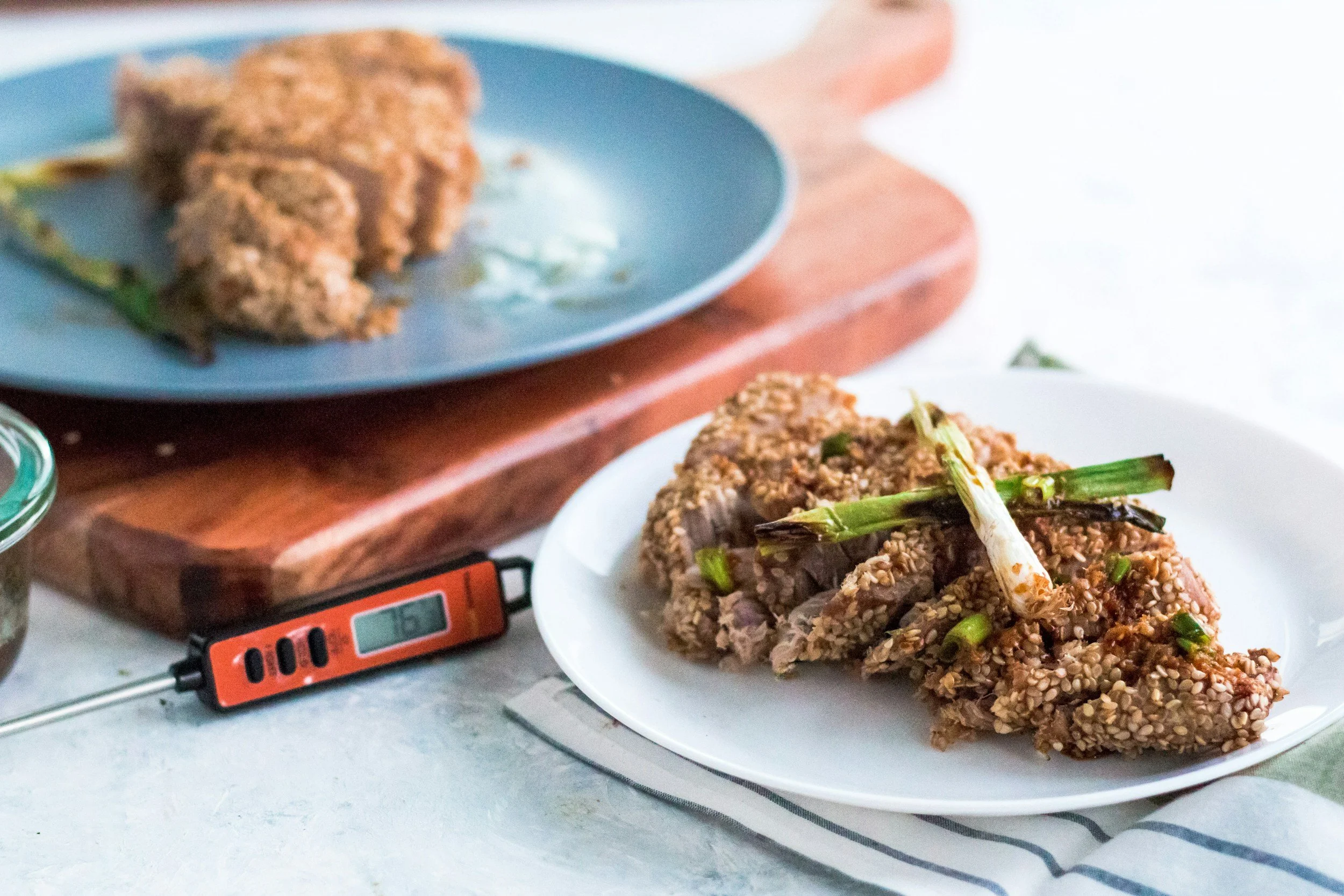 Raw meat coated with sesame seeds on a white plate, with green vegetables on top, placed on a digital kitchen scale.