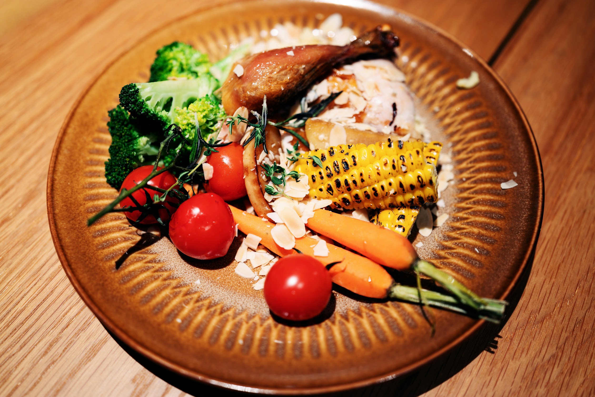 A wooden plate with grilled corn on the cob, cherry tomatoes, broccoli, carrots, a piece of cooked meat, and fresh herbs, garnished with sliced almonds.