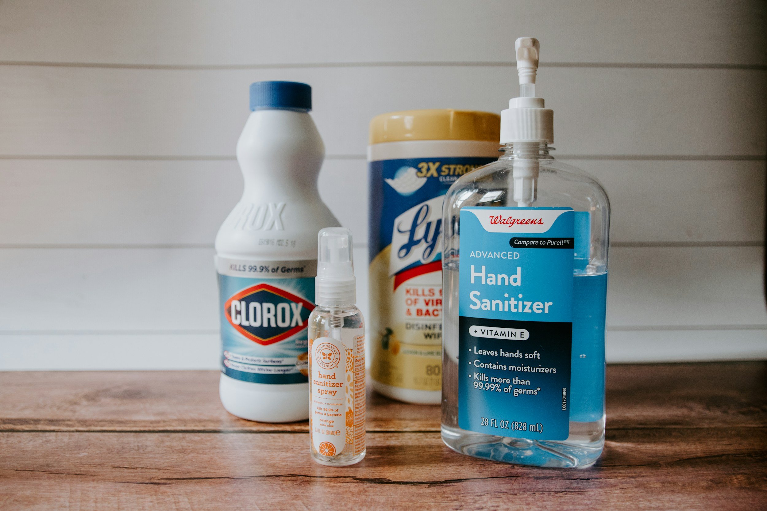 Various disinfectant and sanitizer products on a wooden surface, including a Clorox bleach bottle, a hand sanitizer spray, and a large bottle of Walgreens hand sanitizer.