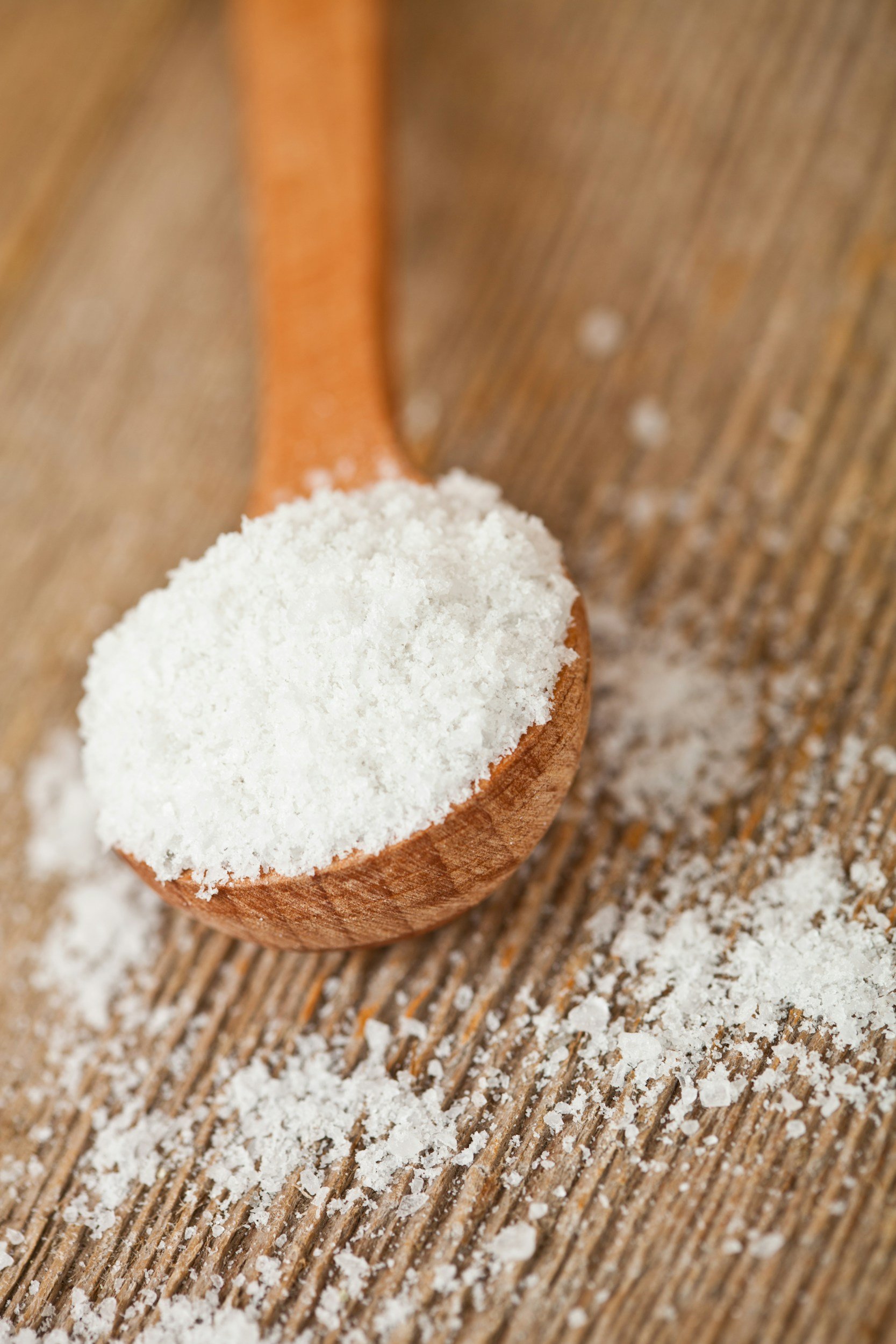 A wooden spoon filled with white flour resting on a wooden surface with scattered flour around.