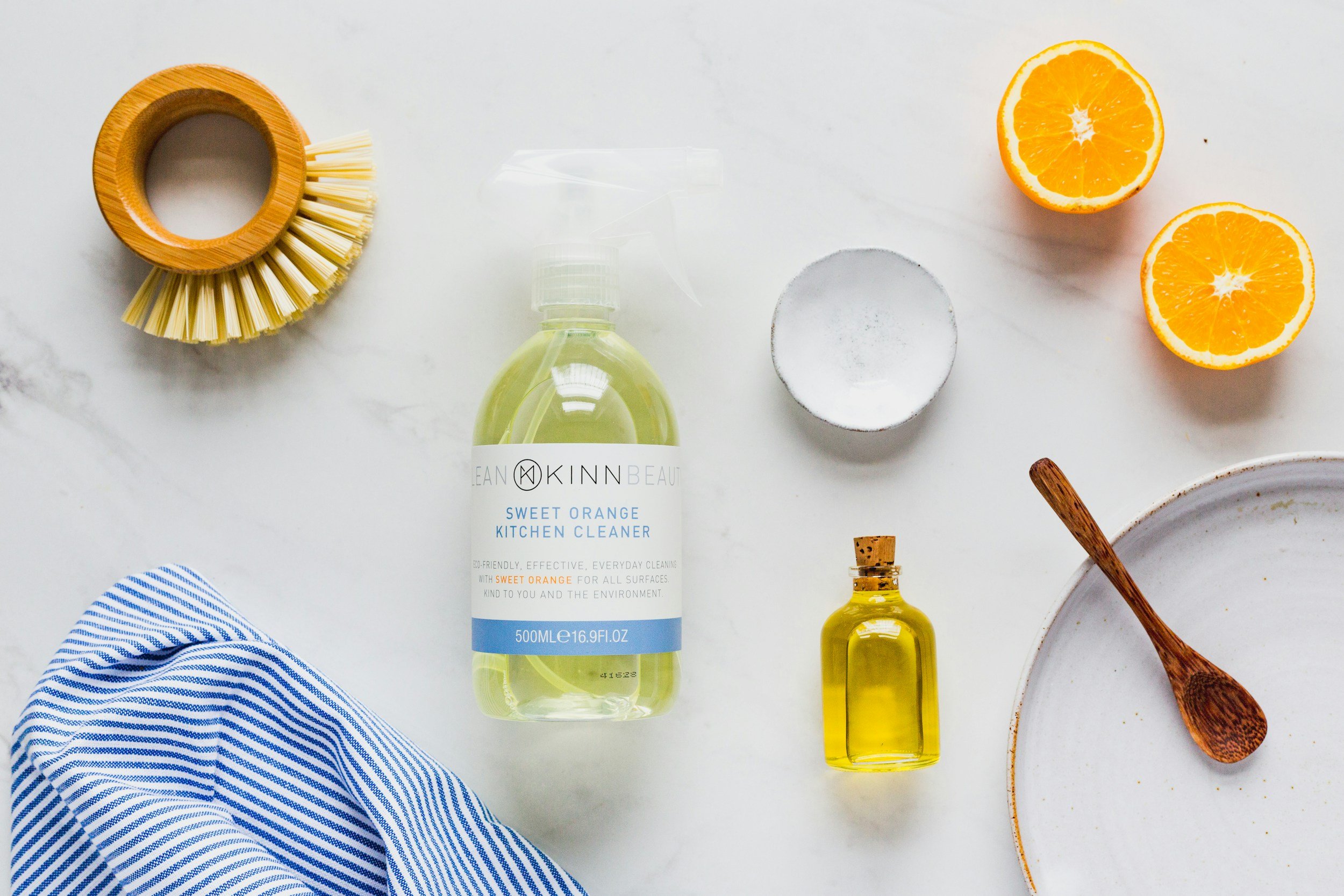 Top view of cleaning supplies and kitchen items on a white surface including a bamboo dish brush, a bottle of sweet orange kitchen cleaner, two halves of an orange, a small bottle of yellow oil, a scrubber, a wooden spoon, part of a striped cloth, and a white bowl.