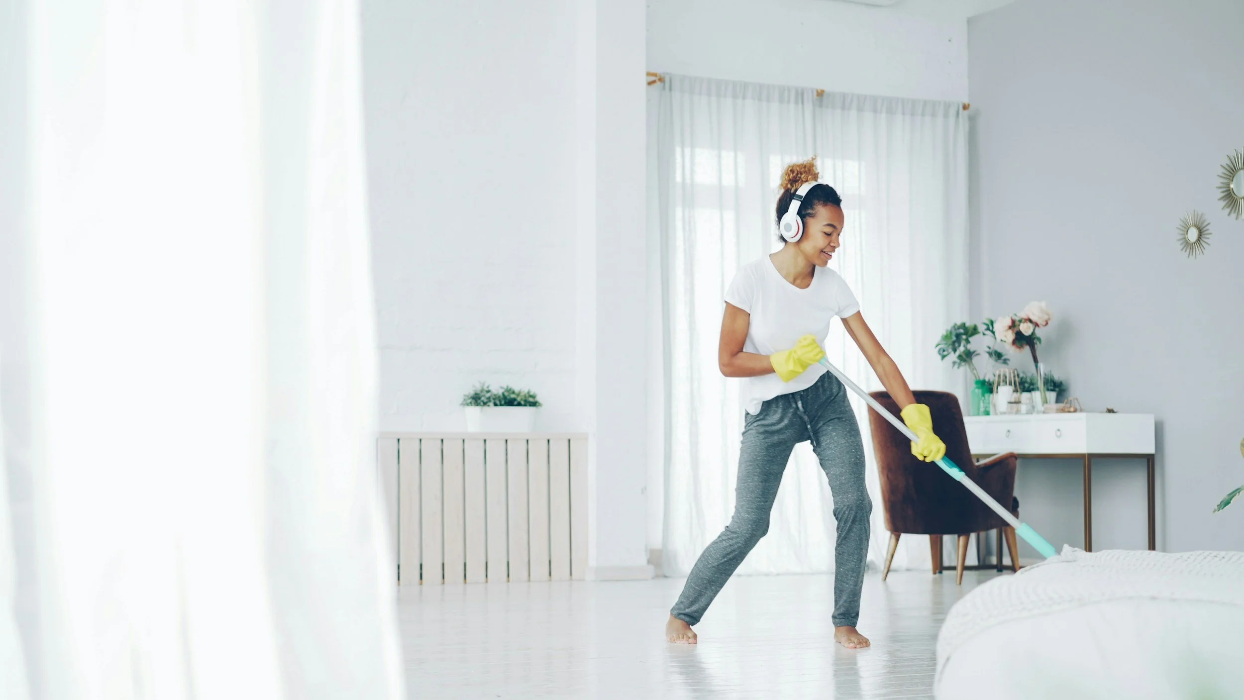 A woman cleaning a bed with a mop in a bright bedroom. She is smiling, wearing yellow gloves, a white T-shirt, and gray sweatpants, with headphones on her head.
