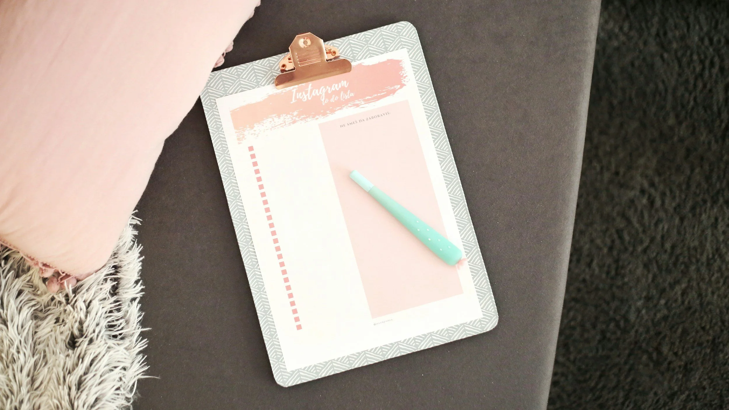 A clipboard with a pink and white sheet of paper attached, with the words 'Instagram' and 'De La Lista' at the top, and a light blue pen placed on the paper, resting on a dark surface next to a fuzzy pink object and a shaggy gray and white textile.
