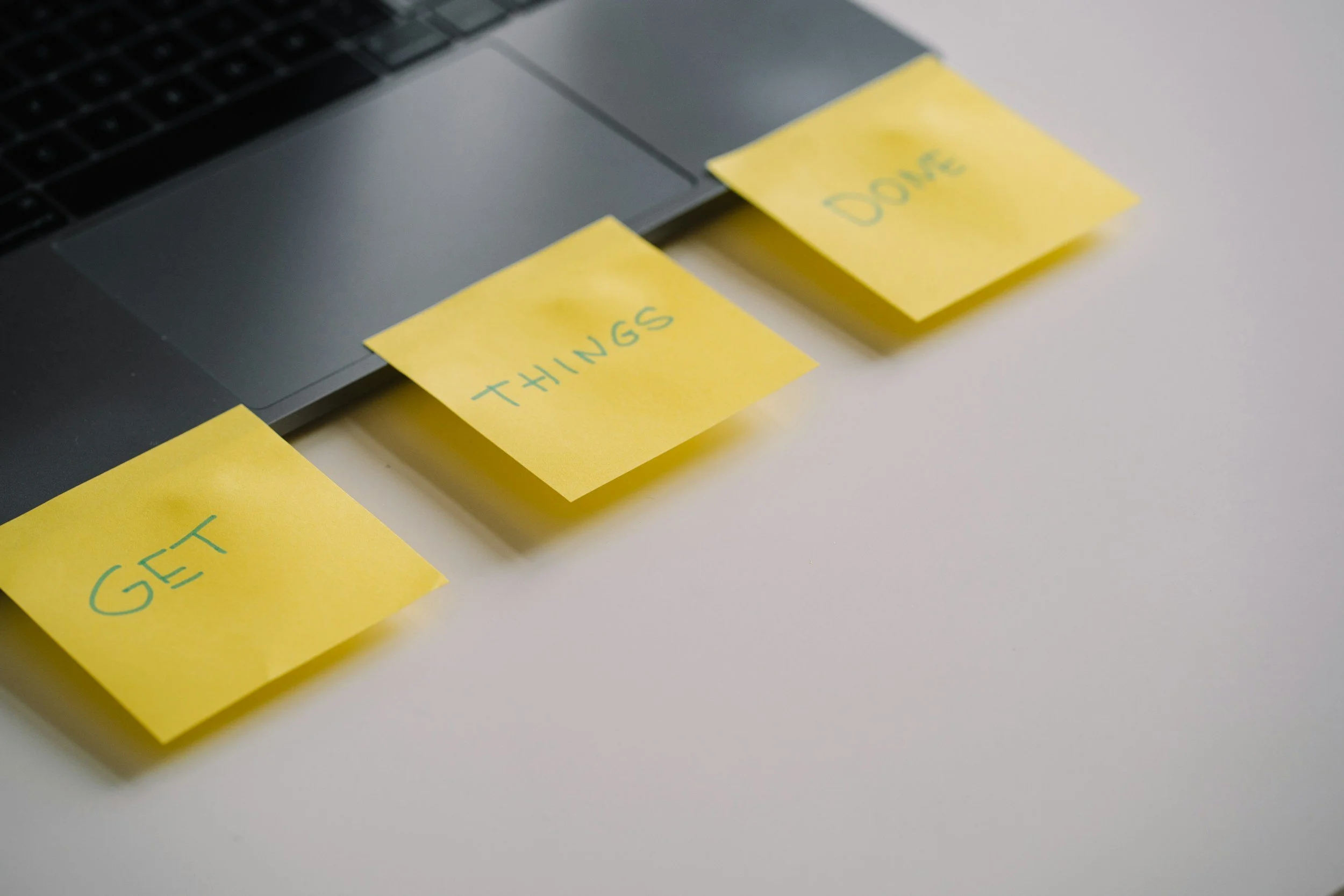 Yellow sticky notes labeled 'GET,' 'THINGS,' and 'DONE' are attached to a gray laptop keyboard on a white surface.