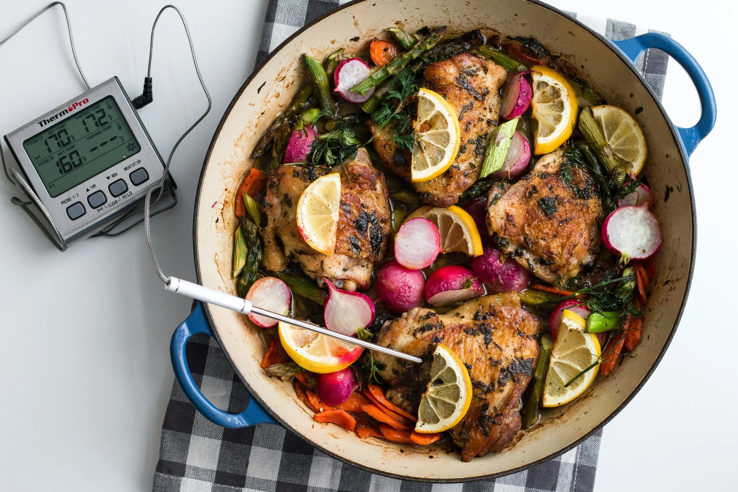 A baked dish in a blue cast iron skillet featuring chicken, lemon slices, radishes, and cooked vegetables including green beans and carrots.