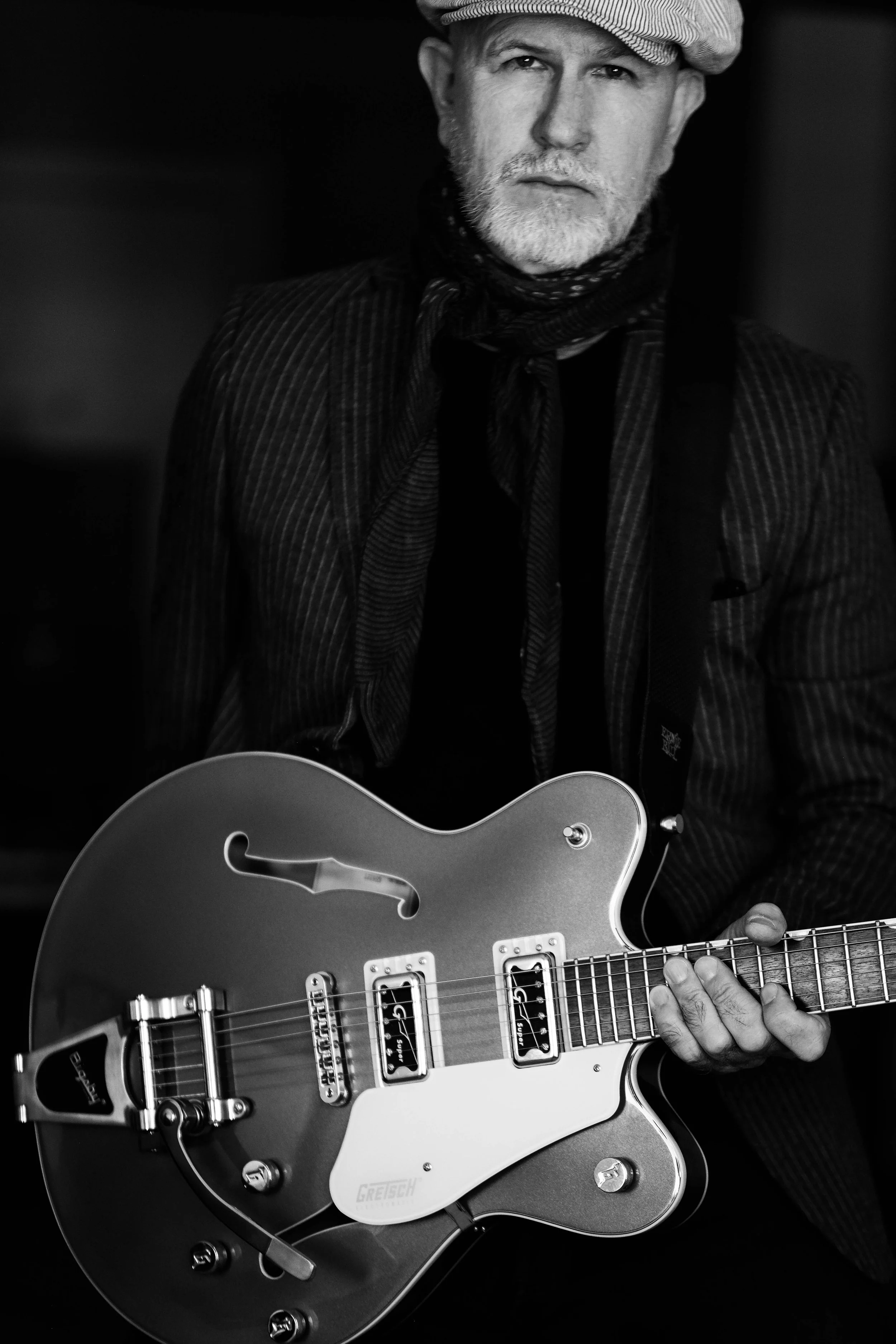 Black and white photo of a songer-songwriter, Christopher Andrew Cooke of the Thrift Store Dandys, wearing a striped jacket, scarf, and newsboy cap, holding a Gretsch electric guitar.