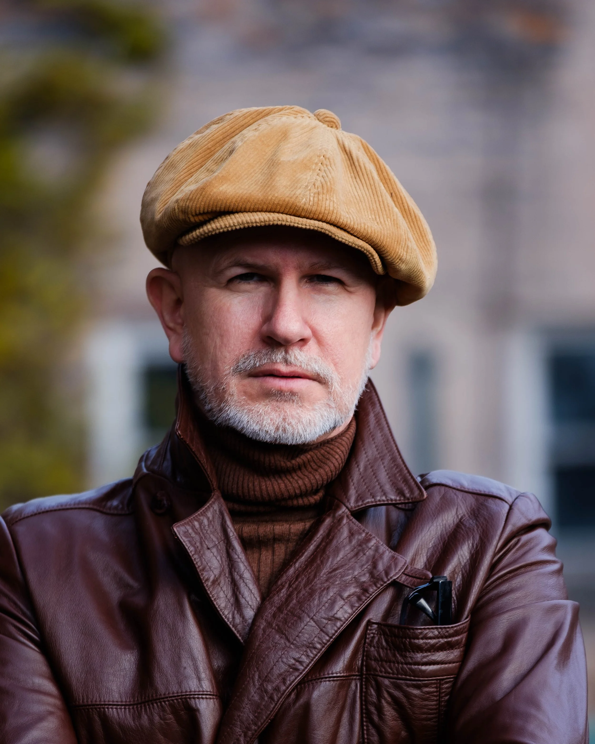 A middle-aged man with a gray beard and mustache wearing a tan newsboy cap, a brown leather jacket, and a dark turtleneck sweater, standing outdoors with a blurred background of trees and buildings.