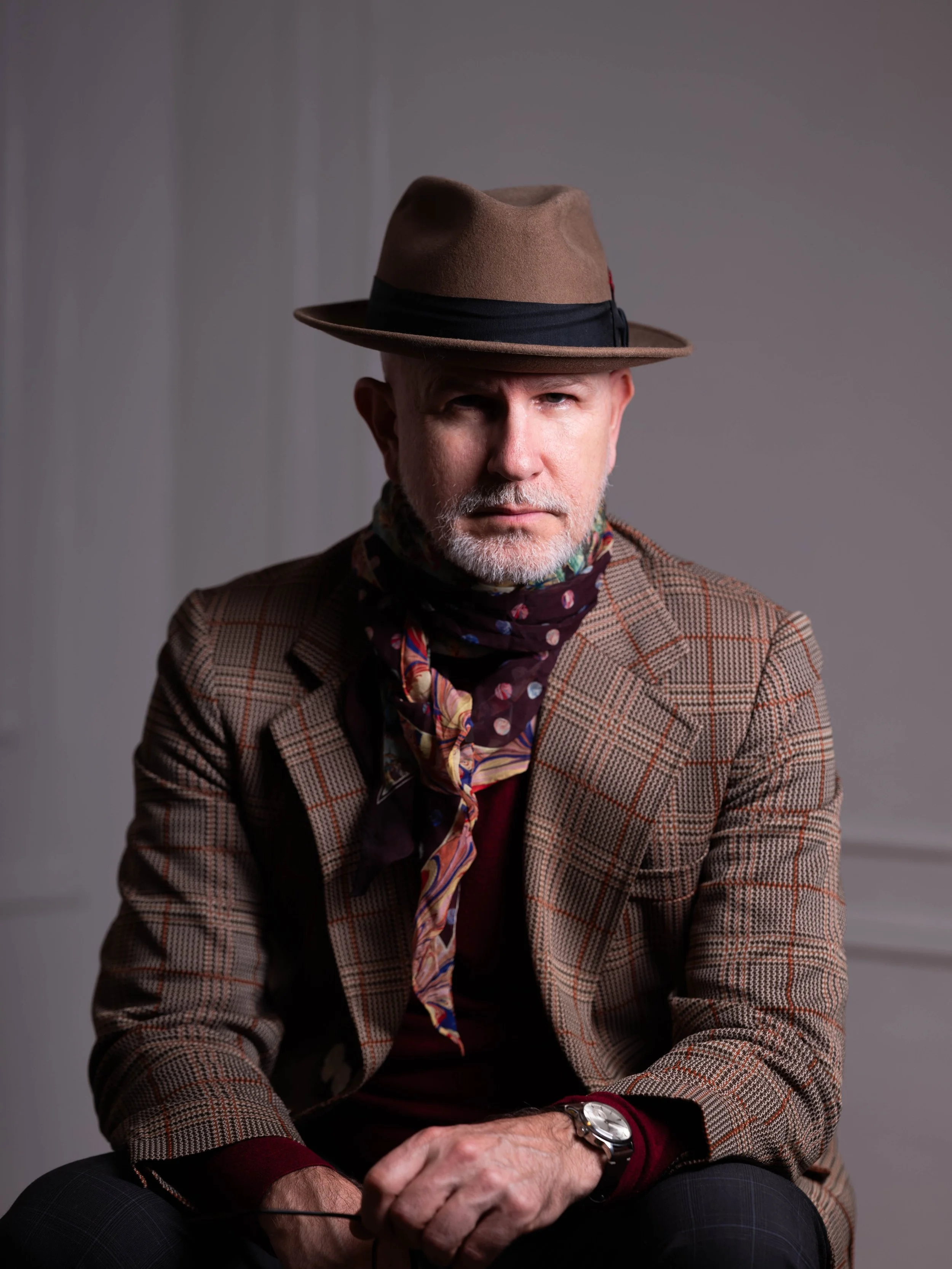 A middle-aged man with a beard wearing a brown hat, a checkered jacket, a colorful neck scarf, and a watch, sitting in front of a gray background.
