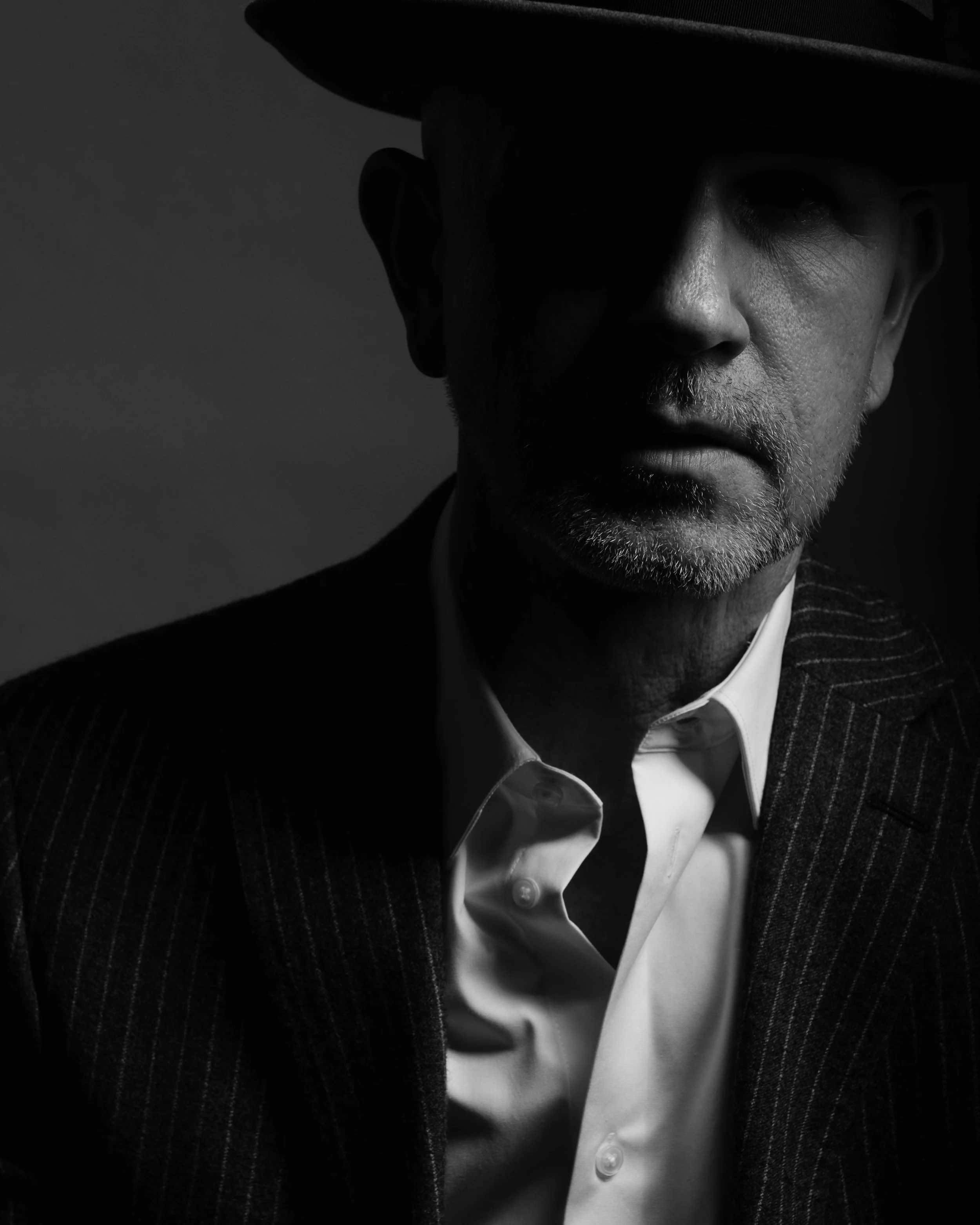 A black-and-white portrait of a man wearing a striped suit, white shirt, and a hat, with dramatic lighting highlighting part of his face.