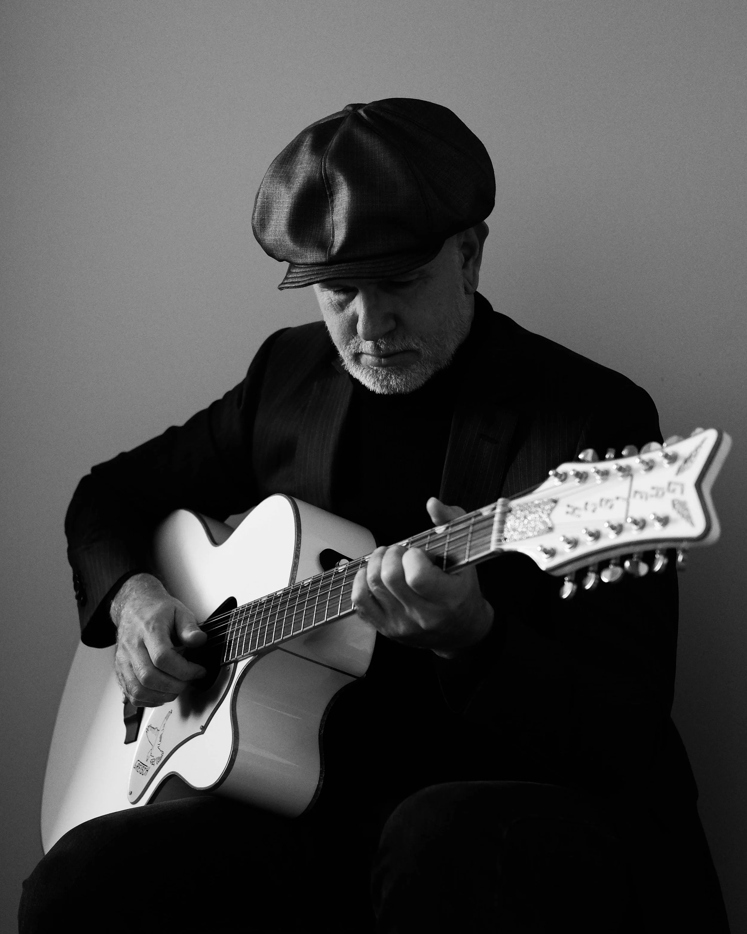 A man wearing a hat and dark clothing, playing an acoustic guitar, pictured in black and white against a plain background.