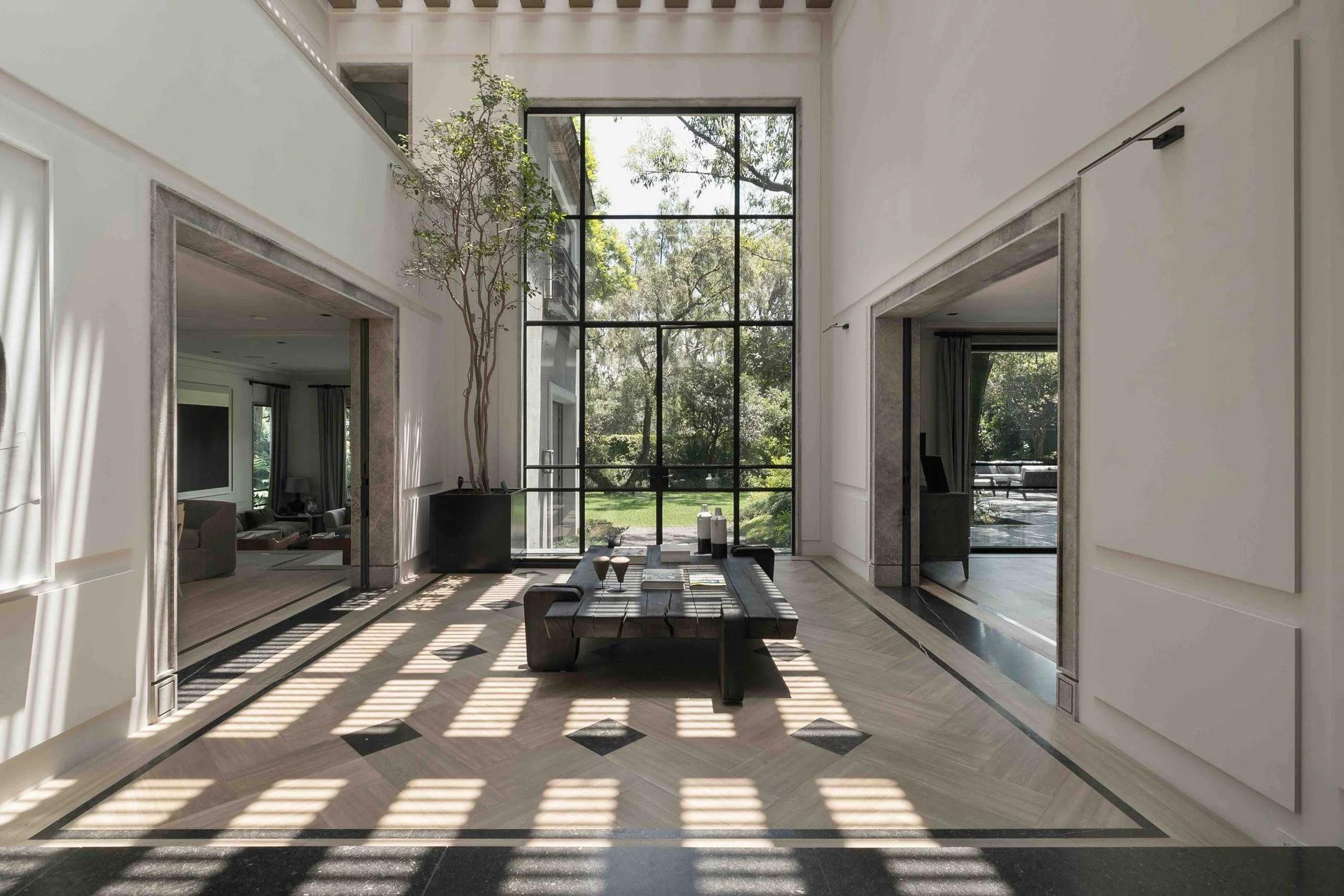 Modern spacious living room with large floor-to-ceiling window overlooking greenery, minimal furniture, and natural sunlight casting shadows on the floor.