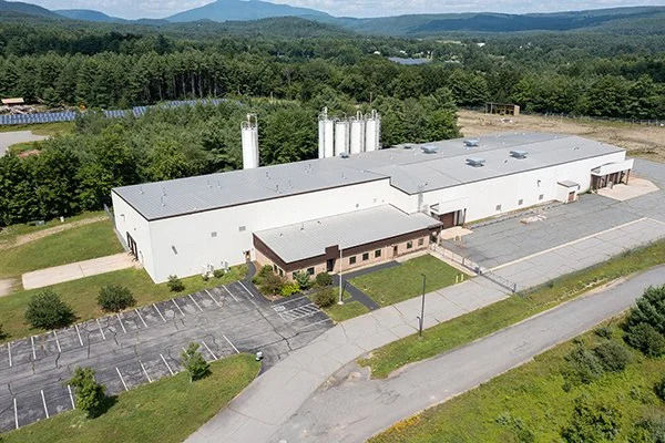 An aerial view of a large white industrial building with parking lots and surrounding green landscape, set against a backdrop of trees and mountains.