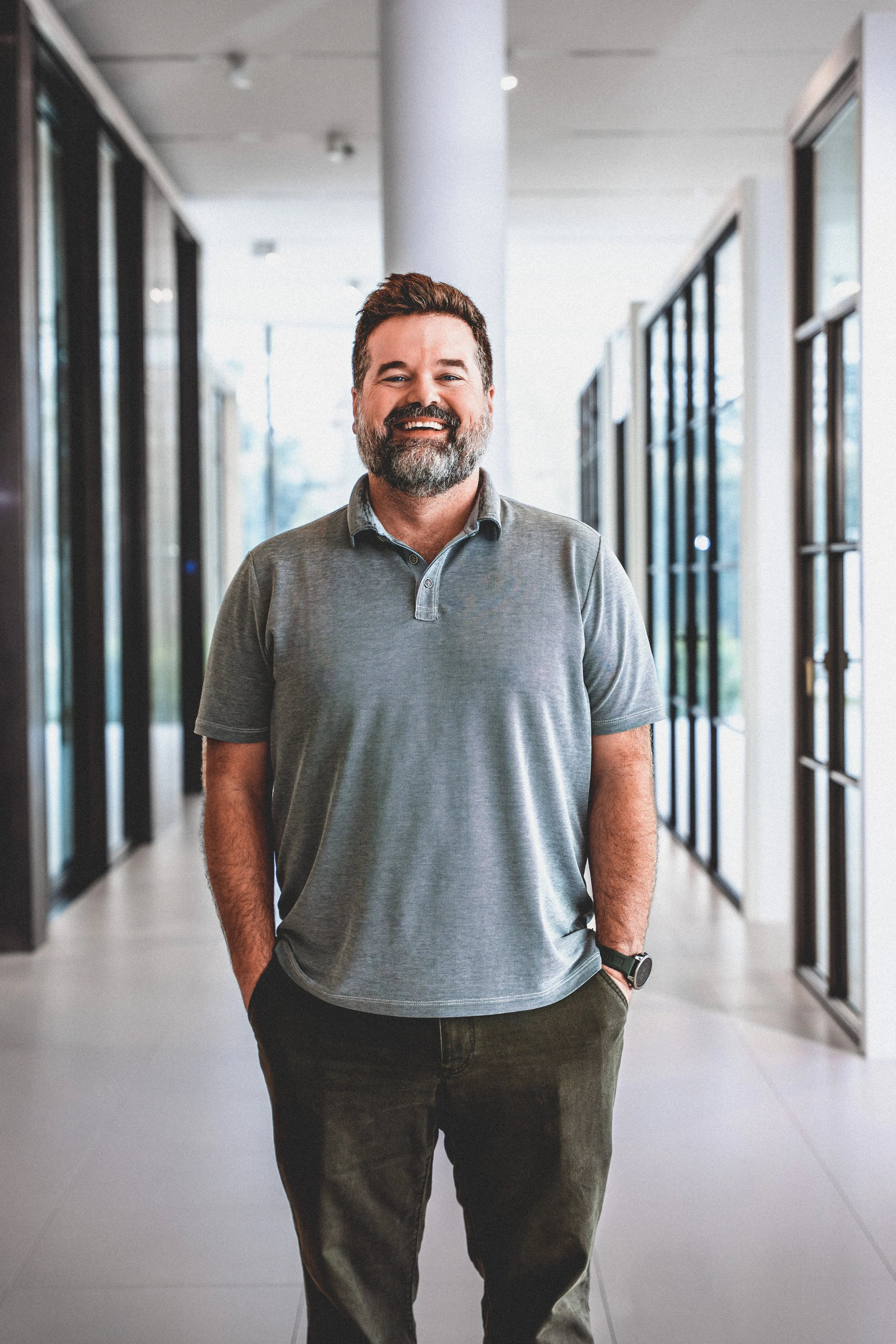 A smiling man with a beard, wearing a gray polo shirt and dark pants, standing in a modern office hallway with glass doors on either side.