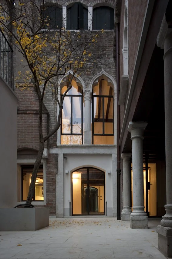 A historic brick building with arched windows reflects the sunset, and a courtyard with a leafless tree is illuminated by warm interior lights.