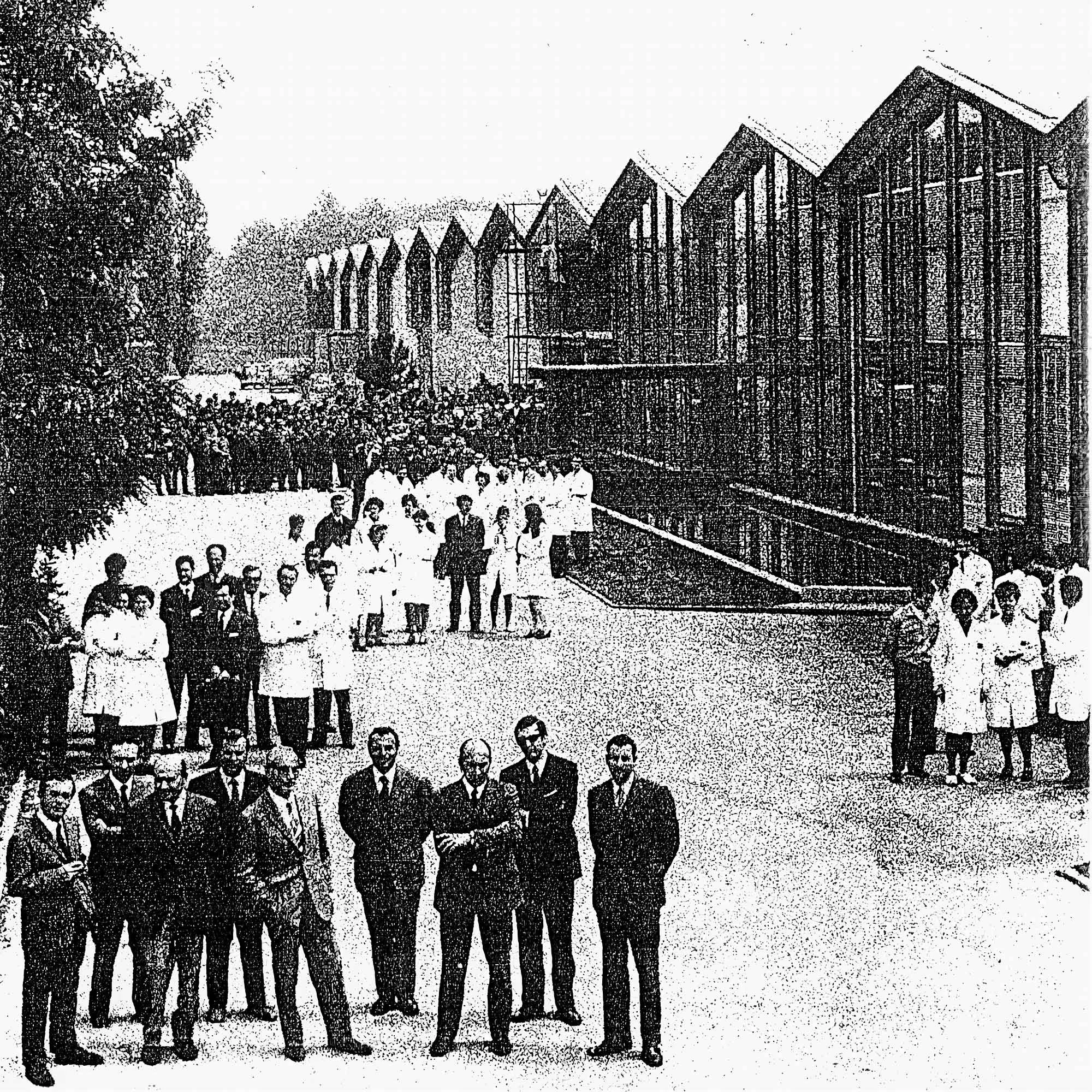 Historical black and white photograph of a large group of men, women, and children gathered outside modern multi-story buildings with large windows, some dressed in medical attire, with trees on the left side.