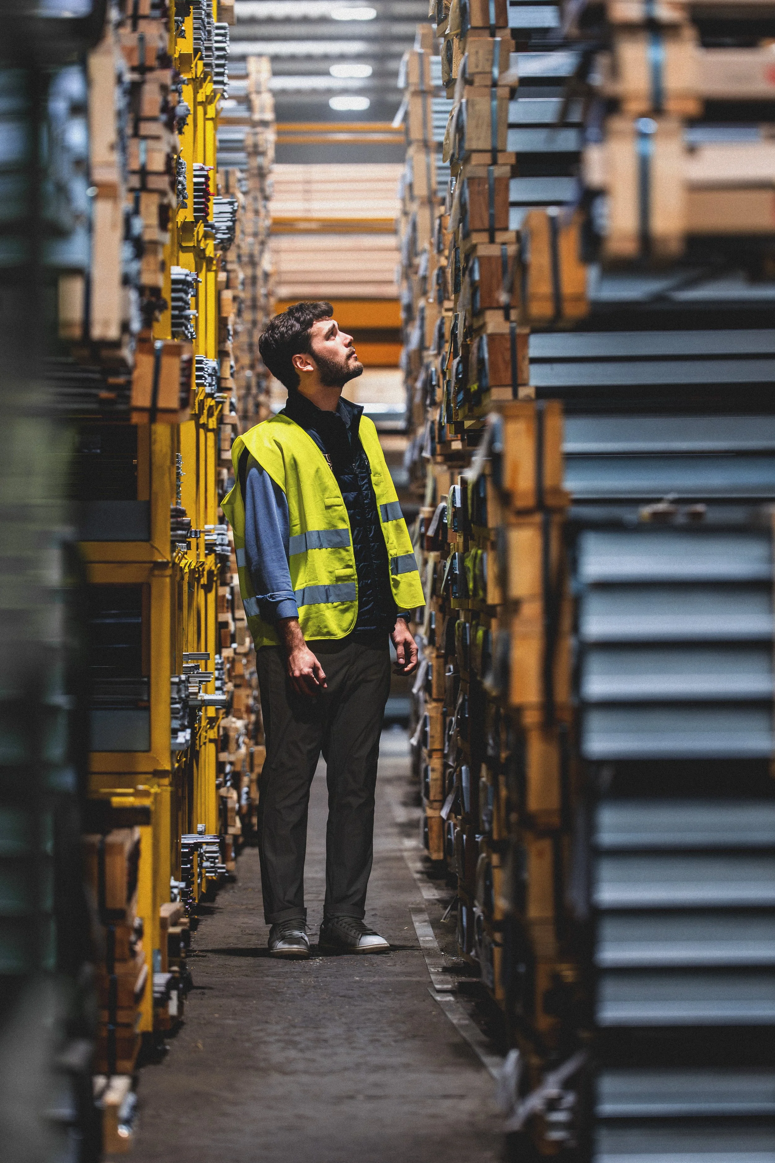 A man in a yellow safety vest standing in a warehouse aisle, surrounded by tall stacks of materials.