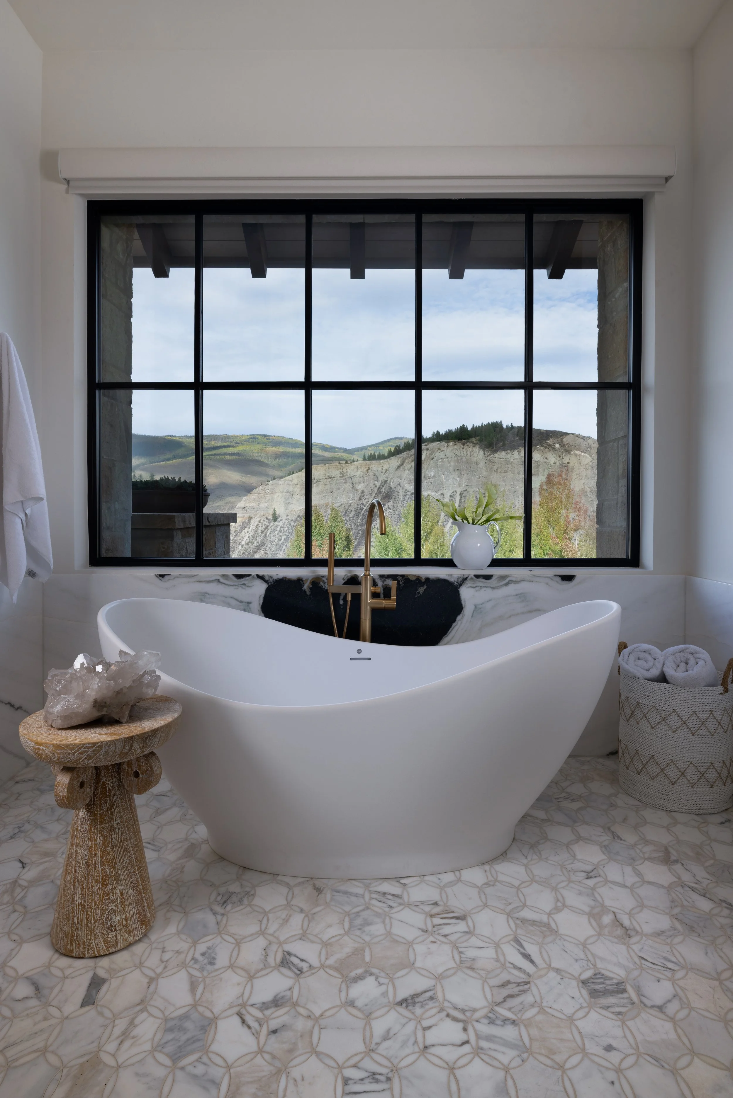 A modern bathroom with a freestanding bathtub large window showing a scenic landscape of hills and trees. There is a small wooden stool with a crystal on top, a white vase with green leaves, a basket with rolled towels, and a white towel hanging on the wall.