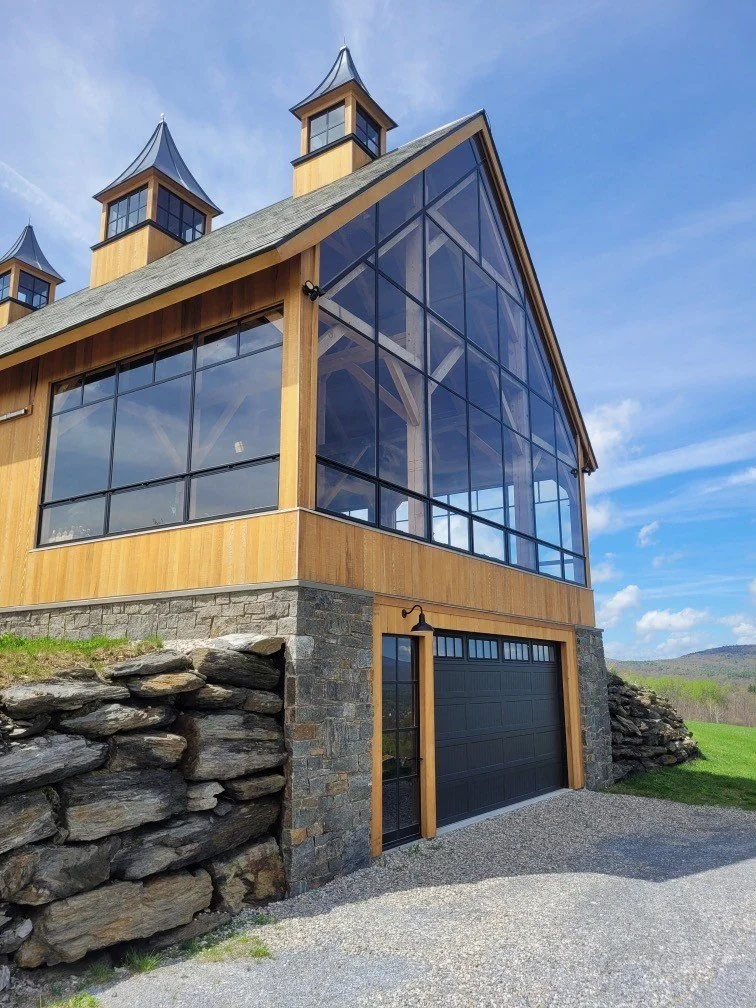 A modern barn-style building with large custom steel windows, wooden siding, stone foundation, and small tower structures on the roof, set against a blue sky with some clouds.