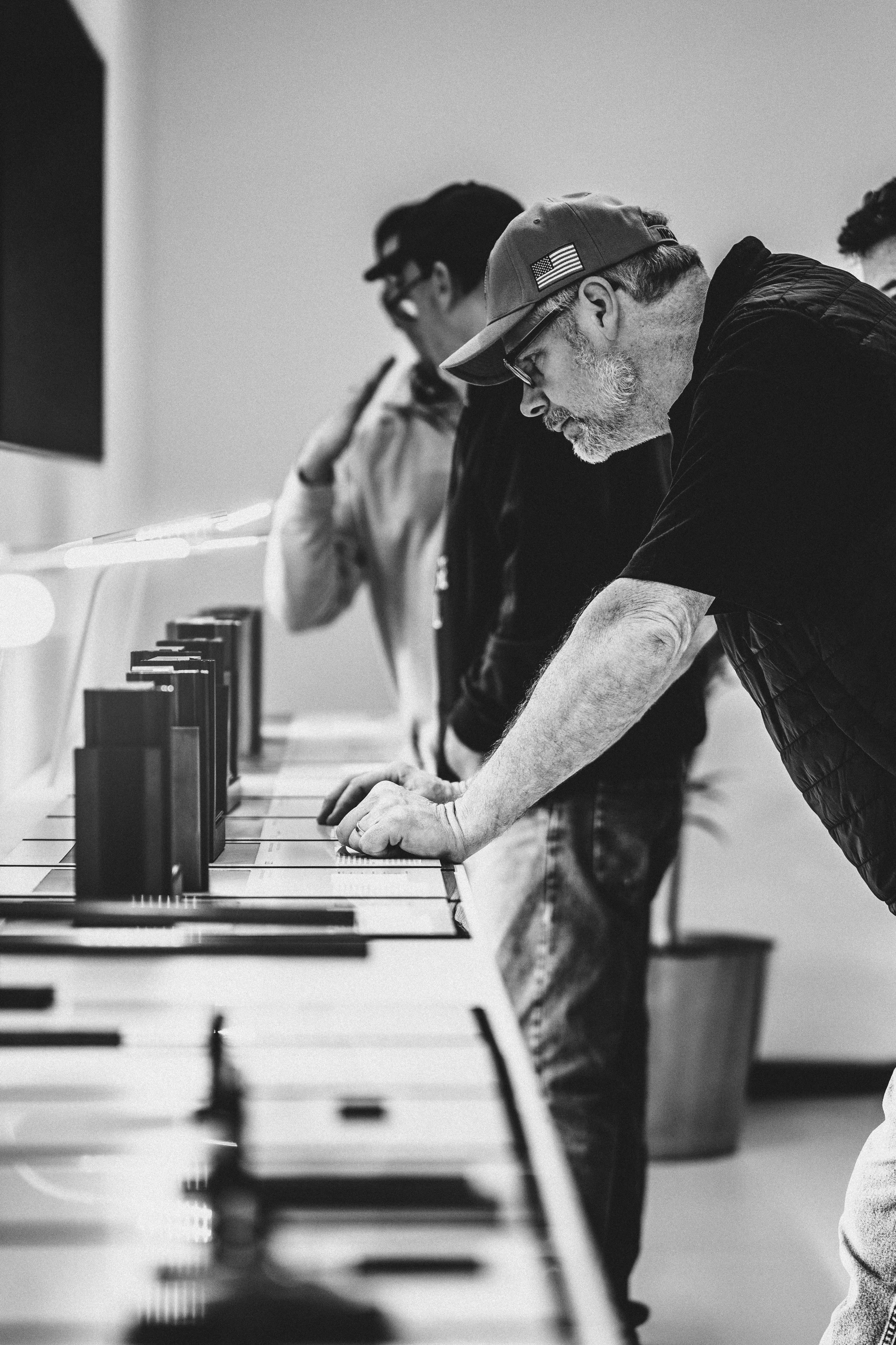A black and white photo of a man leaning over a piano, concentrating on playing. He wears a cap with an American flag patch and glasses. In the background, another person is also focused on the piano.