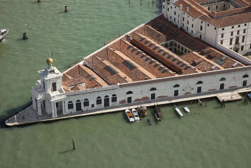 Aerial view of a historic building in Venice surrounded by water, with boats docked nearby, and red-tiled roofs.