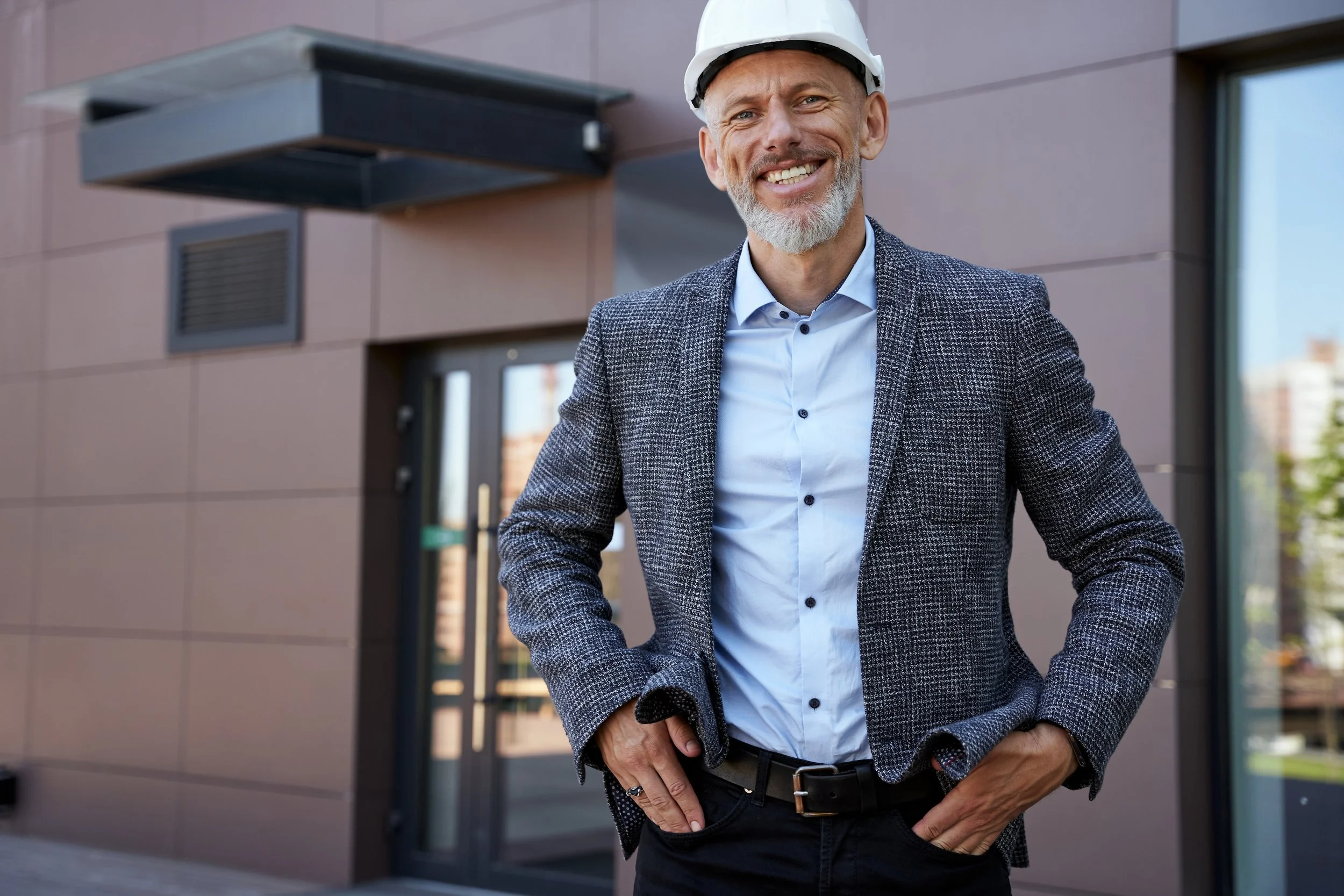 A smiling middle-aged man wearing a white safety helmet and a checkered blazer standing outside a modern building.