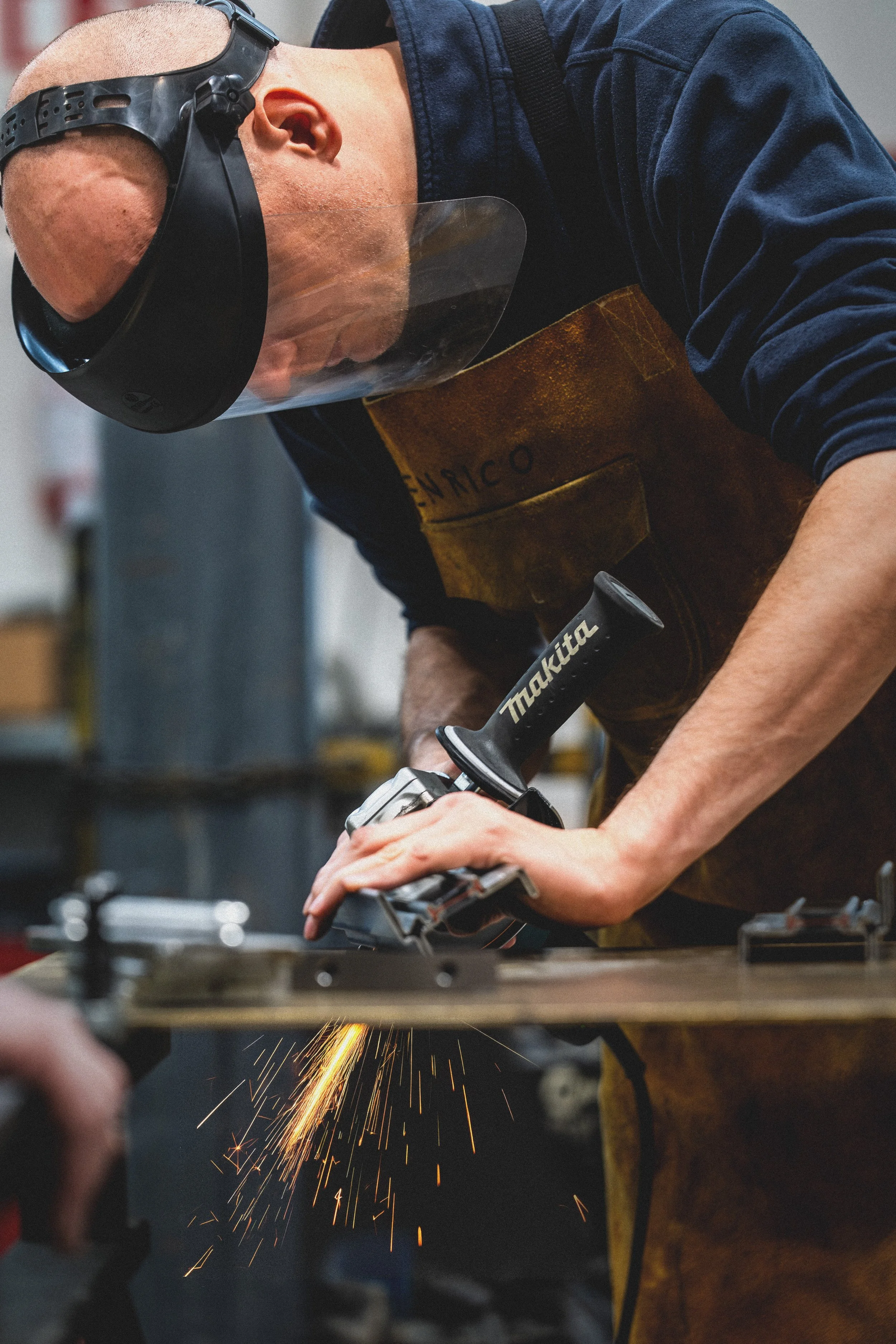 A man wearing safety goggles and a brown apron is using a power tool to cut metal, with sparks flying from the cut.