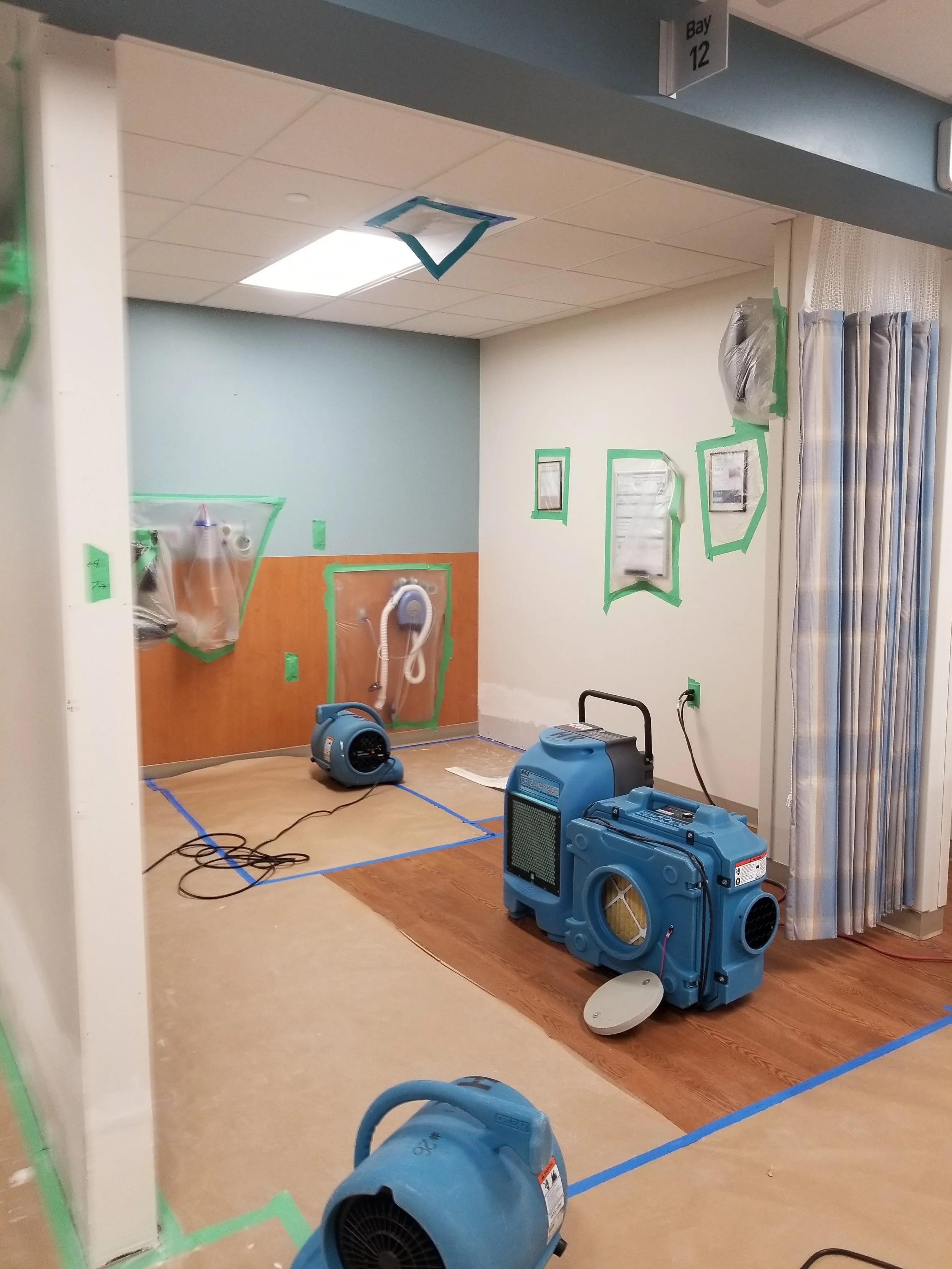 Room under renovation with drying equipment, plastic-covered objects taped to the wall, and a sign indicating Bay 12.
