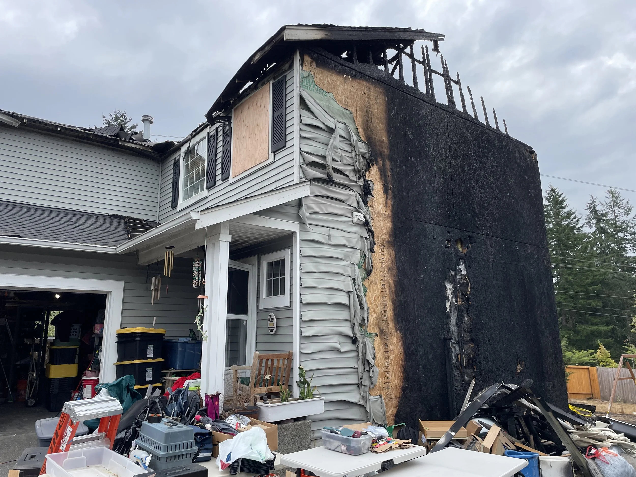 A house with significant fire damage on one side, showing blackened, charred construction and a missing roof section. The other side of the house appears intact with various household items and clutter in the driveway and porch area.