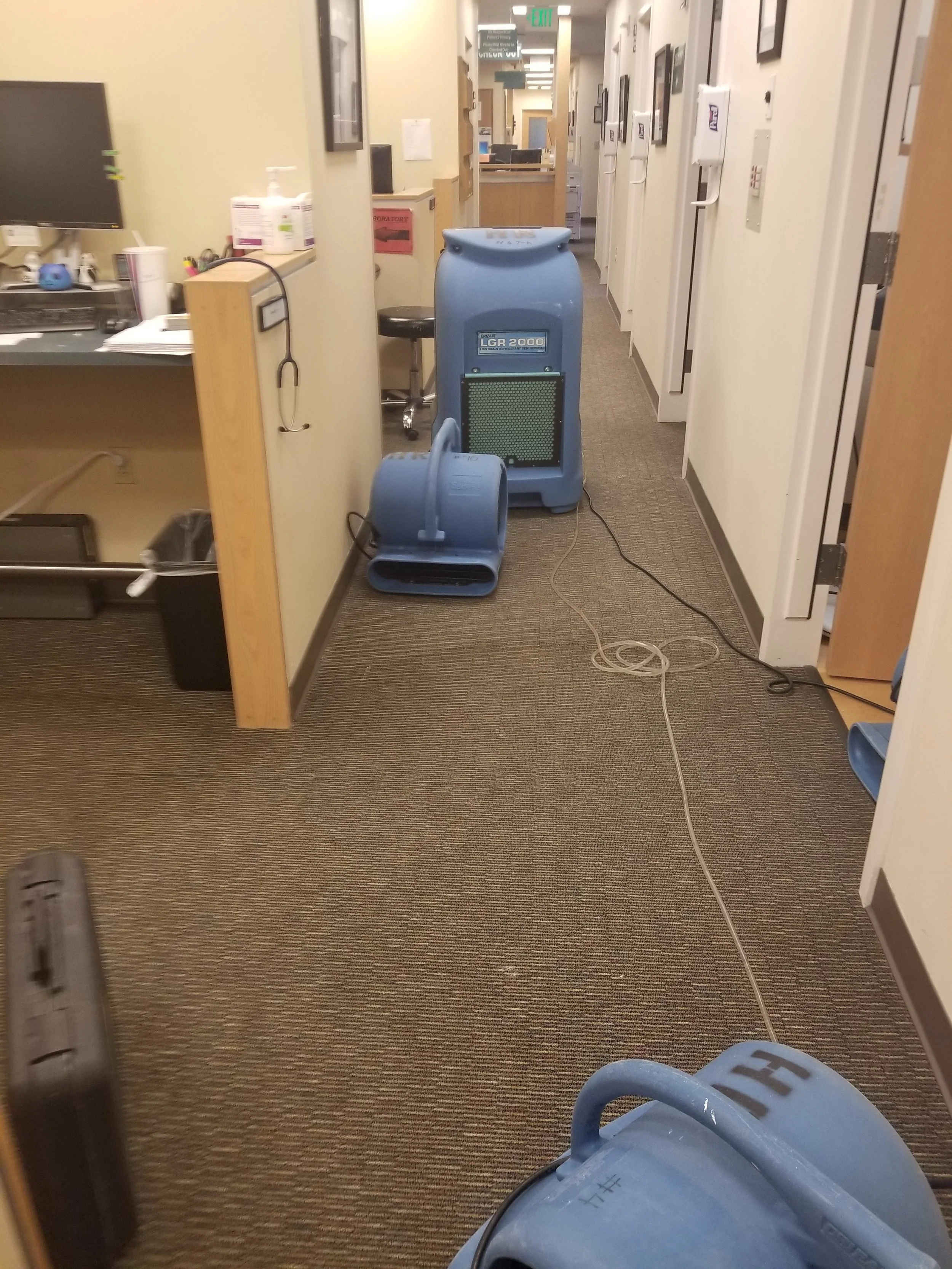Hospital corridor with medical equipment, including a large blue medical device on wheels, a ventillator, and various supplies on shelves along the wall.