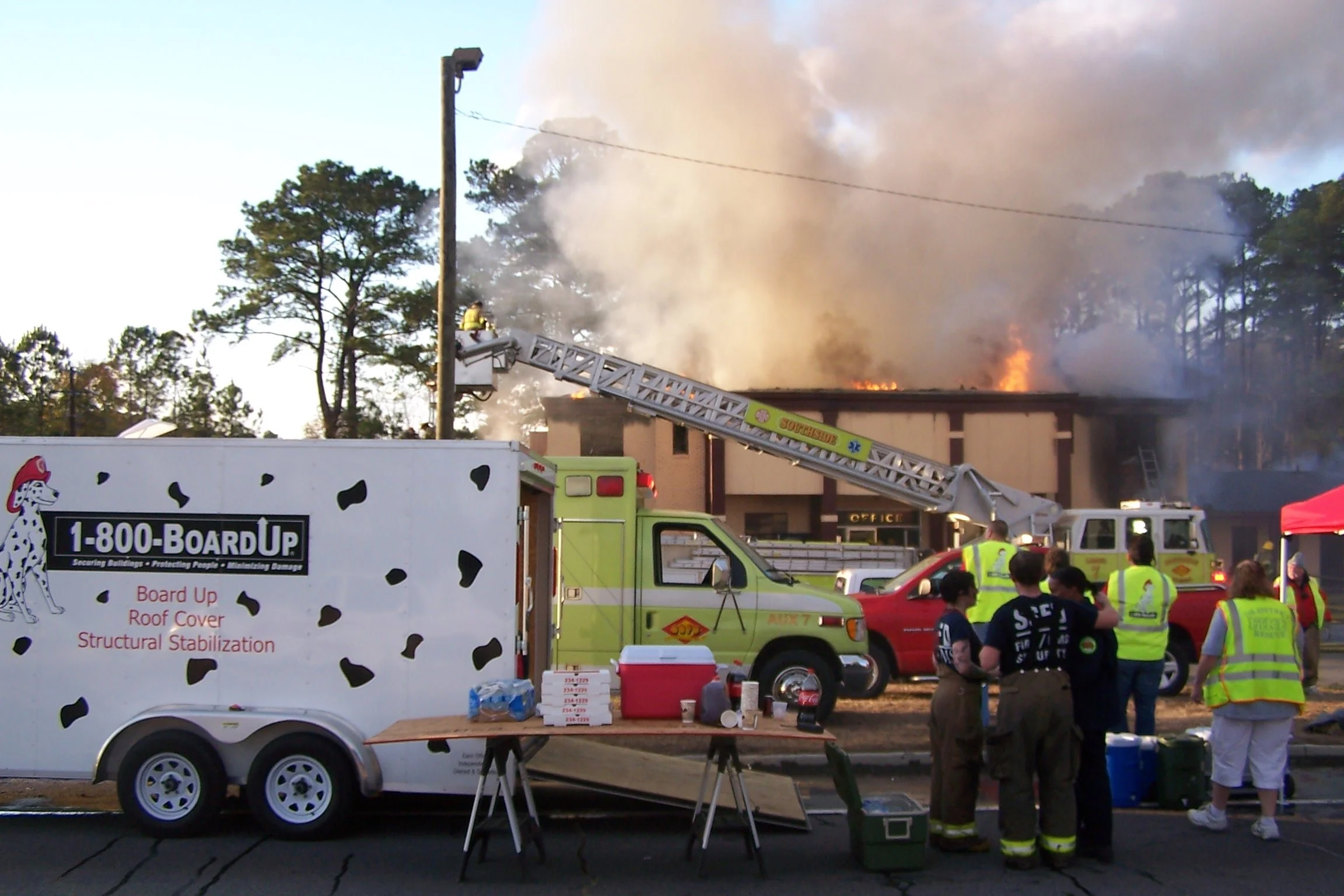 Firefighters combat a building fire with smoke and flames visible, using a ladder truck, as onlookers and emergency vehicles are present.