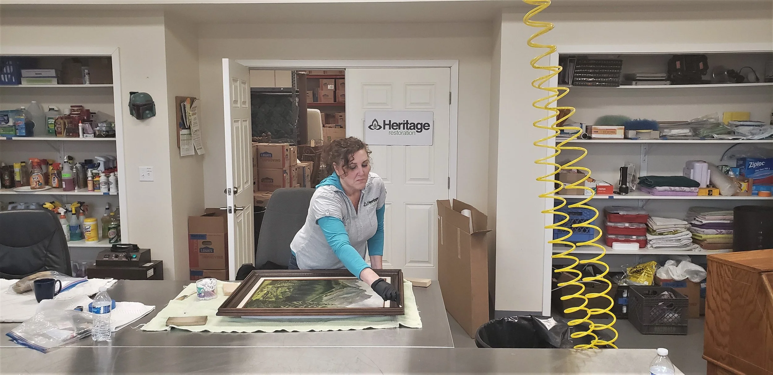 A woman in a gray and turquoise Heritage Restoration uniform handling a framed landscape painting at a work table in a storage or workspace area with shelves, boxes, and cleaning supplies.