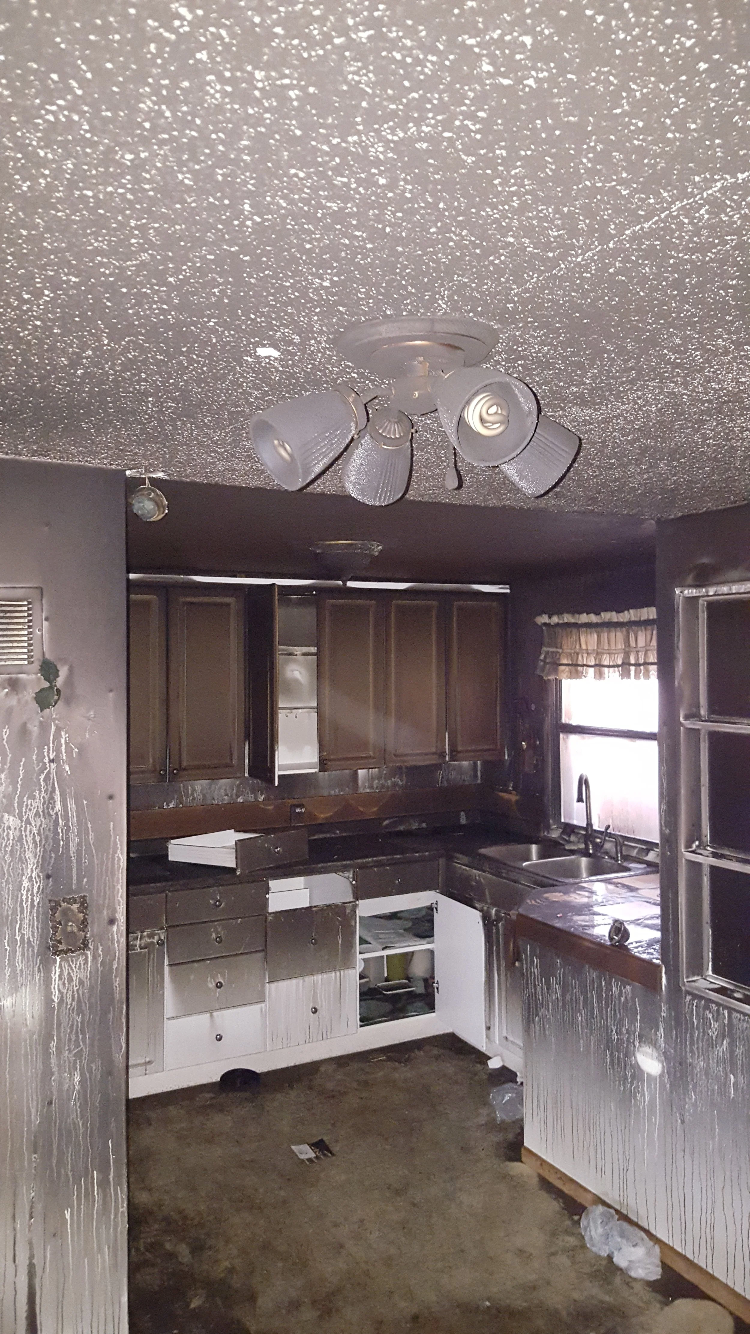 A kitchen damaged by fire, with soot-covered cabinets, walls, and appliances, and a ceiling fan with soot around it.