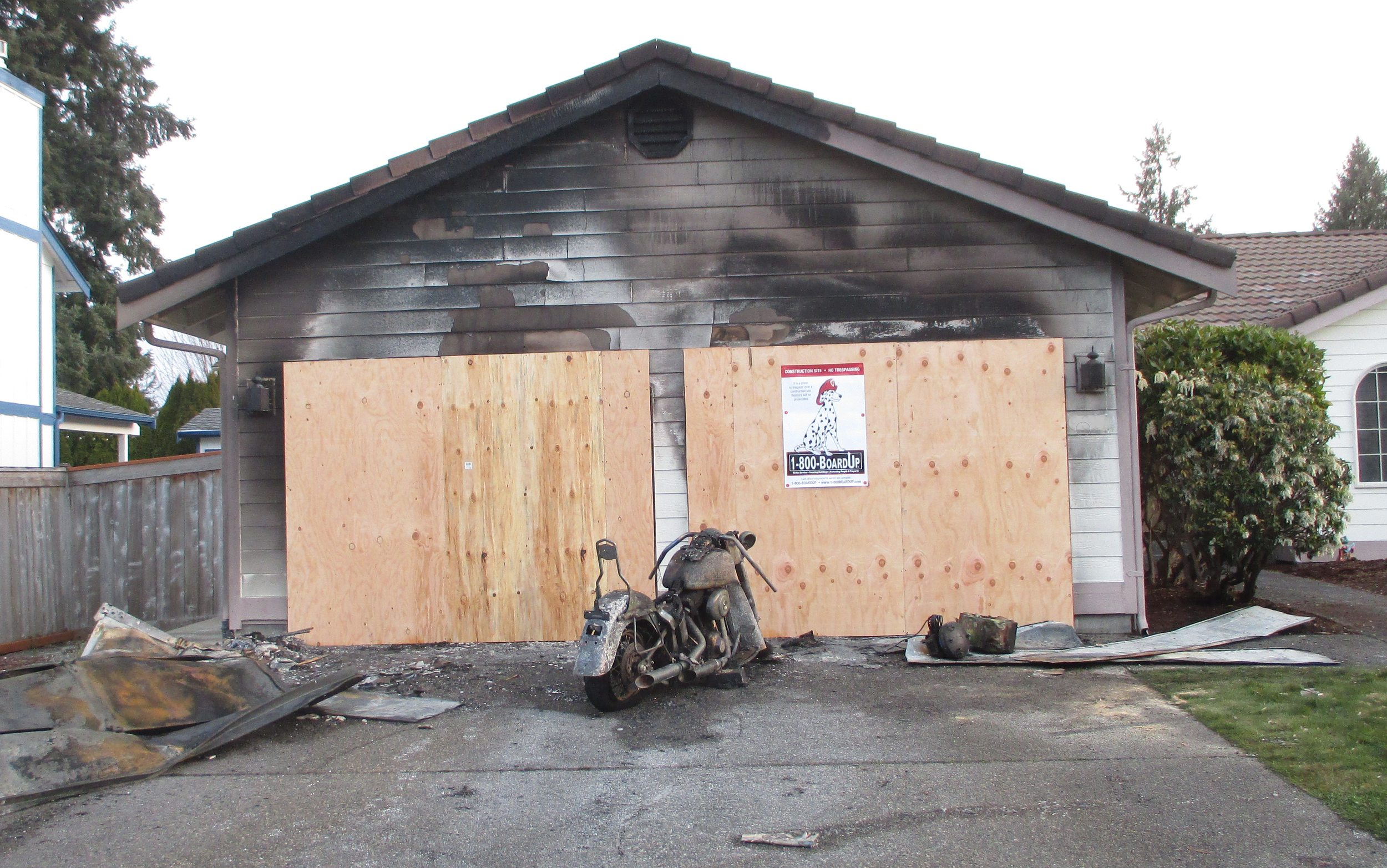A house with blackened and charred exterior walls, plywood boarded-up windows and a blackened motorcycle in front, indicating recent fire damage.