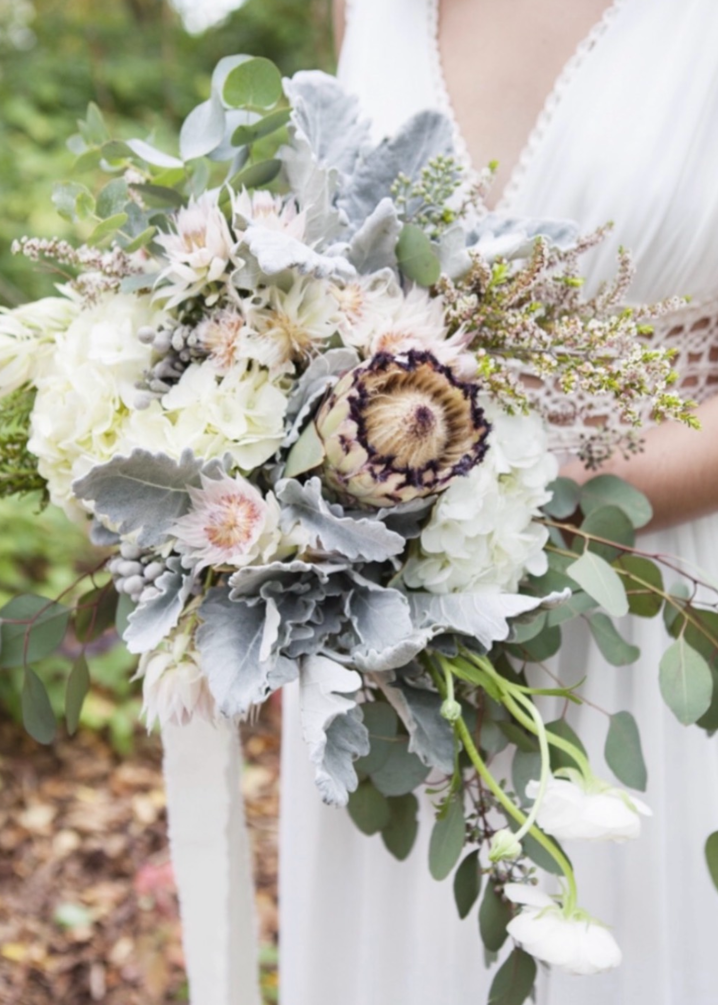 Wedding bouquet featuring white and gray flowers, eucalyptus leaves, and a cotton flower, held by a bride in a white dress.