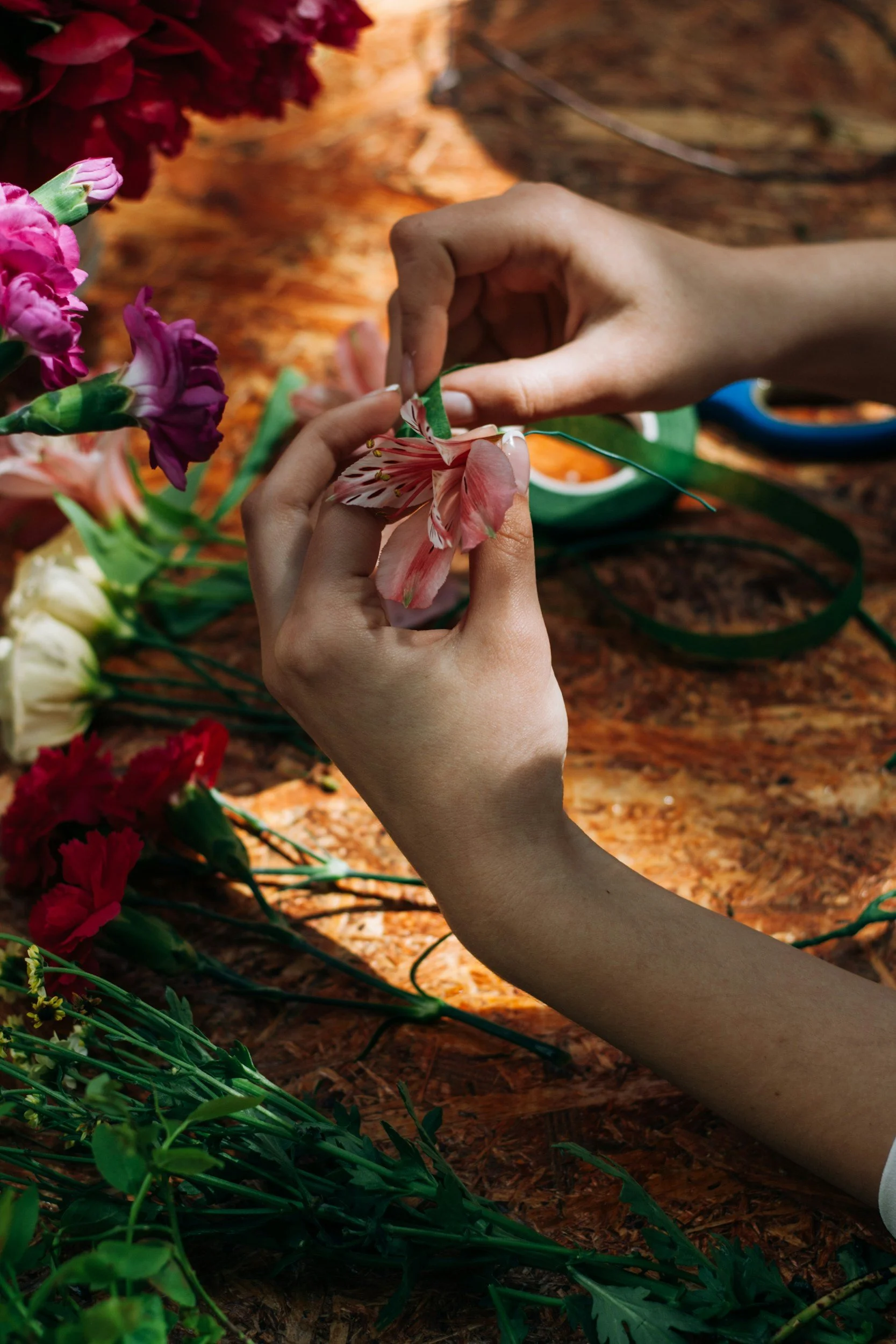 Hands assembling a pink and white flower with floral tape on a dark wooden table, surrounded by various flowers and green floral wire.