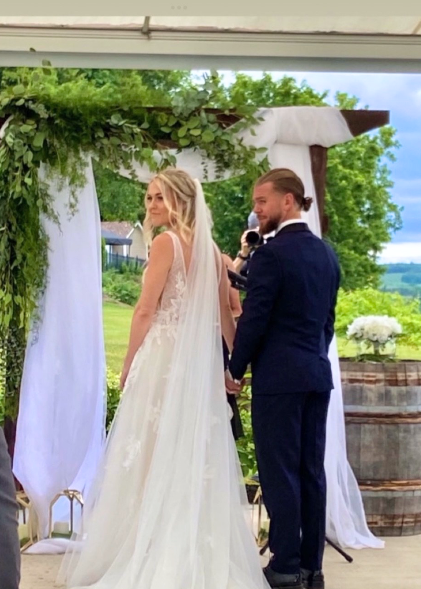 A bride and groom holding hands during their outdoor wedding ceremony, standing under a decorated arch with greenery and white drapery, with a scenic green landscape in the background.